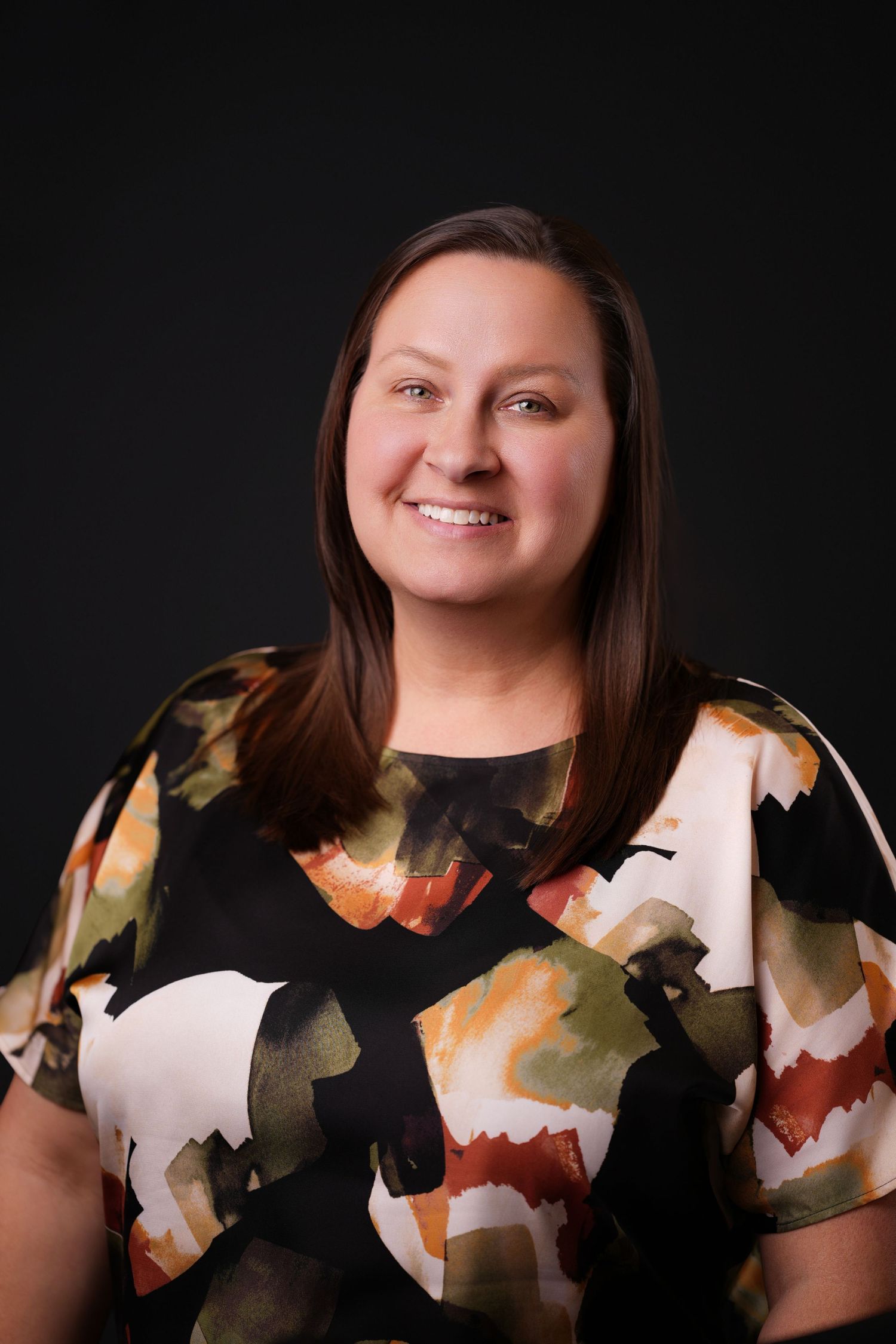 Smiling woman with long brown hair wearing a black blouse with an orange, green, and cream abstract pattern against a dark background.