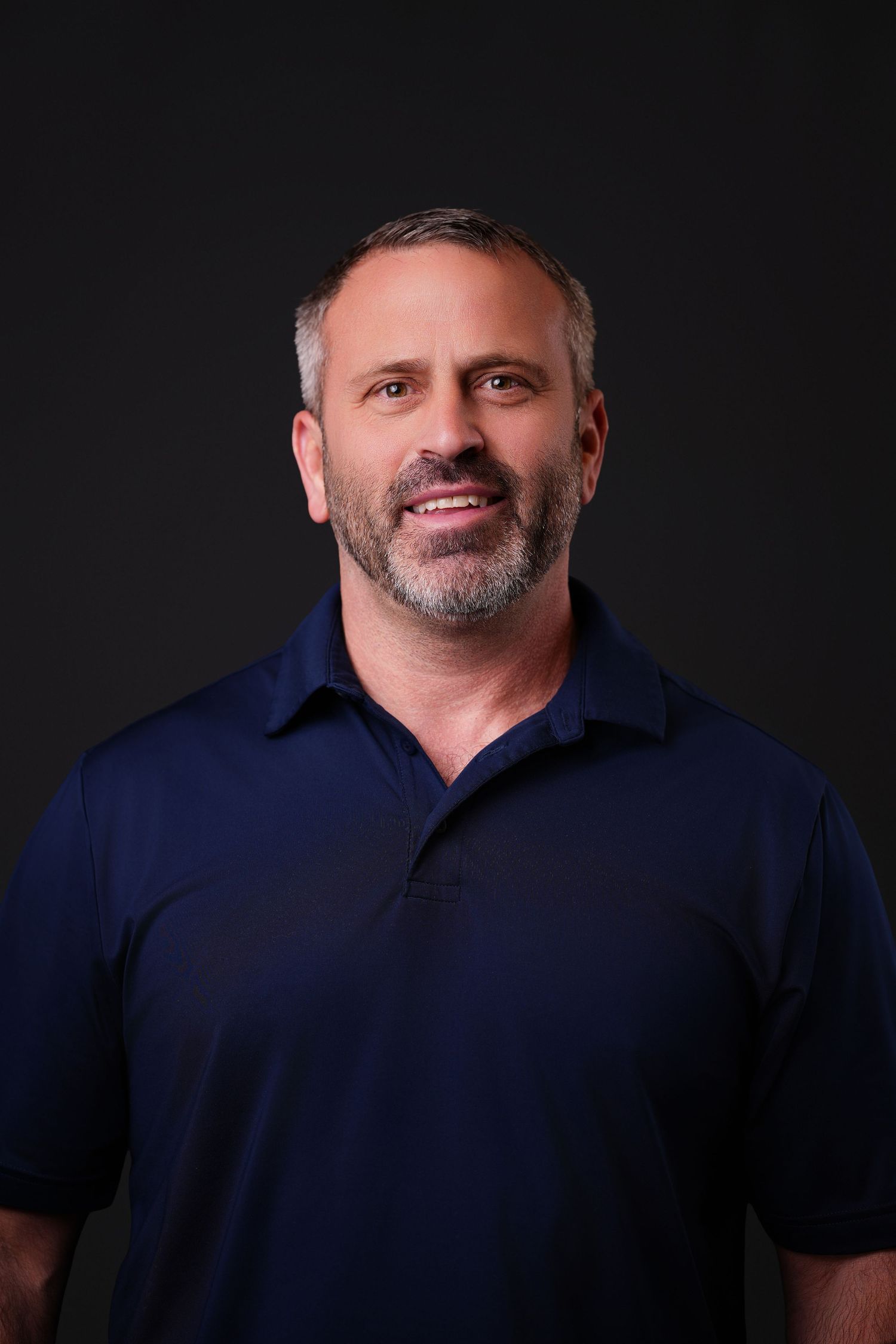 Smiling middle-aged man with short gray hair and a salt-and-pepper beard wearing a navy polo shirt against a dark studio background.