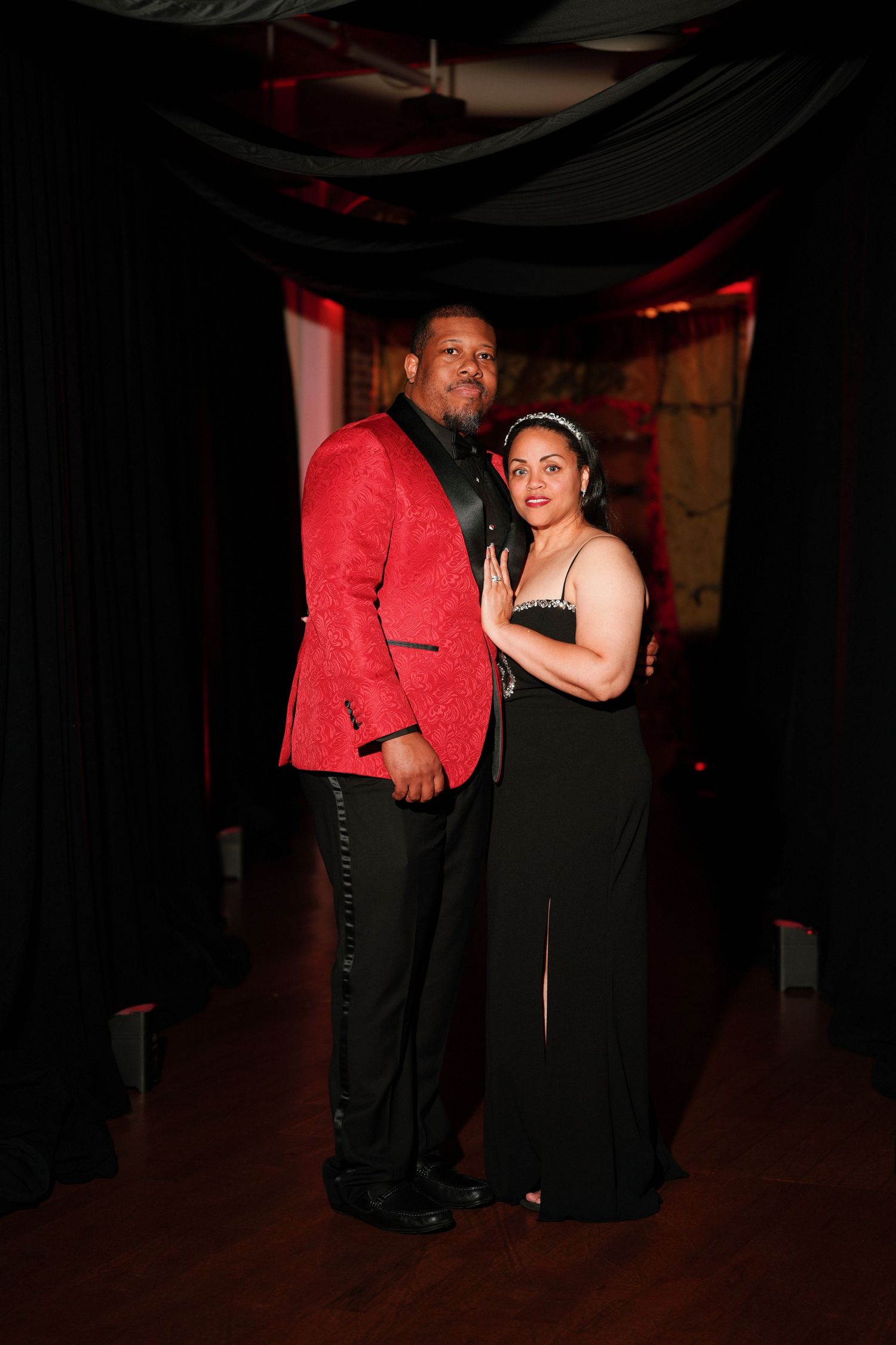Stylish couple photographed at a formal event in Charlotte, NC, featuring a red sequin blazer, black evening gown, and dramatic dark curtain