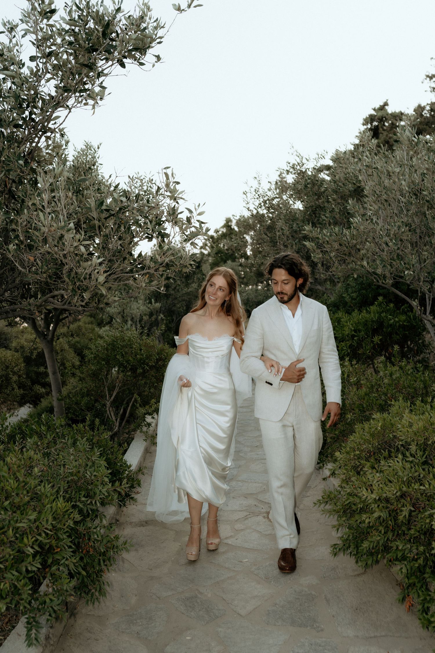 A couple in white attire walks together on a garden path surrounded by lush greenery and trees. Paros Wedding