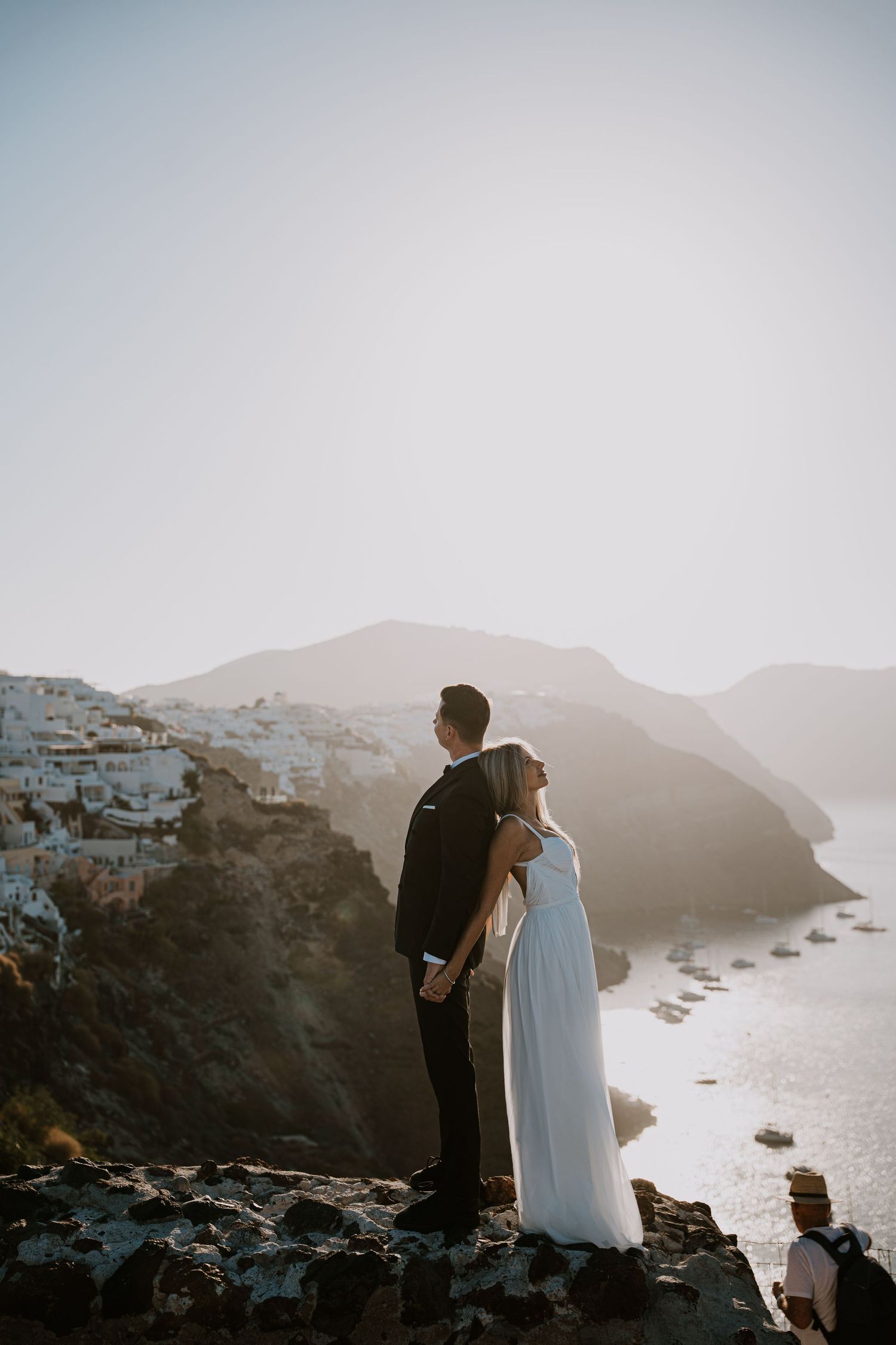 A couple stands on a rocky cliff overlooking a misty coastal landscape during sunset in Santorini, Greece.