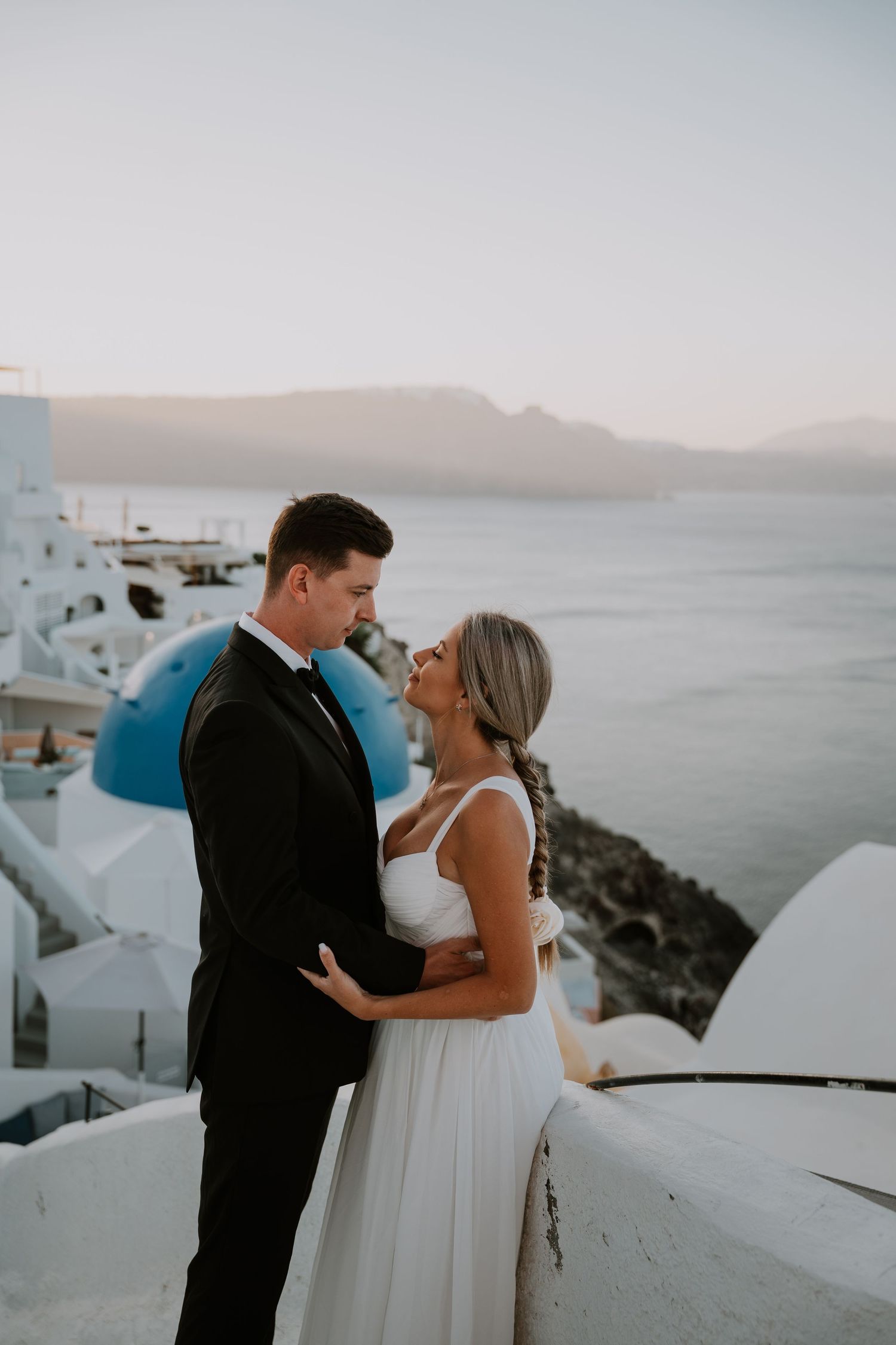 A romantic wedding couple embraces against the backdrop of Santorini's white buildings and blue sea at sunset.