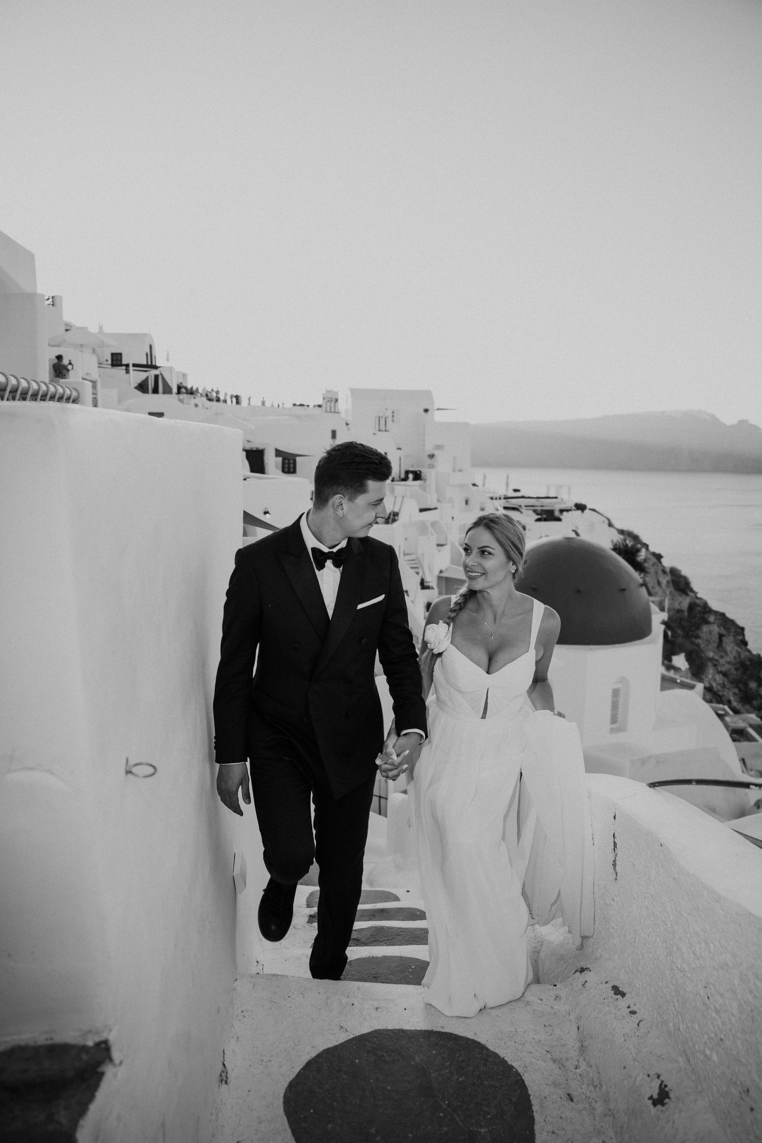 Wedding couple walking hand in hand through Santorini's white pathways with sea views.