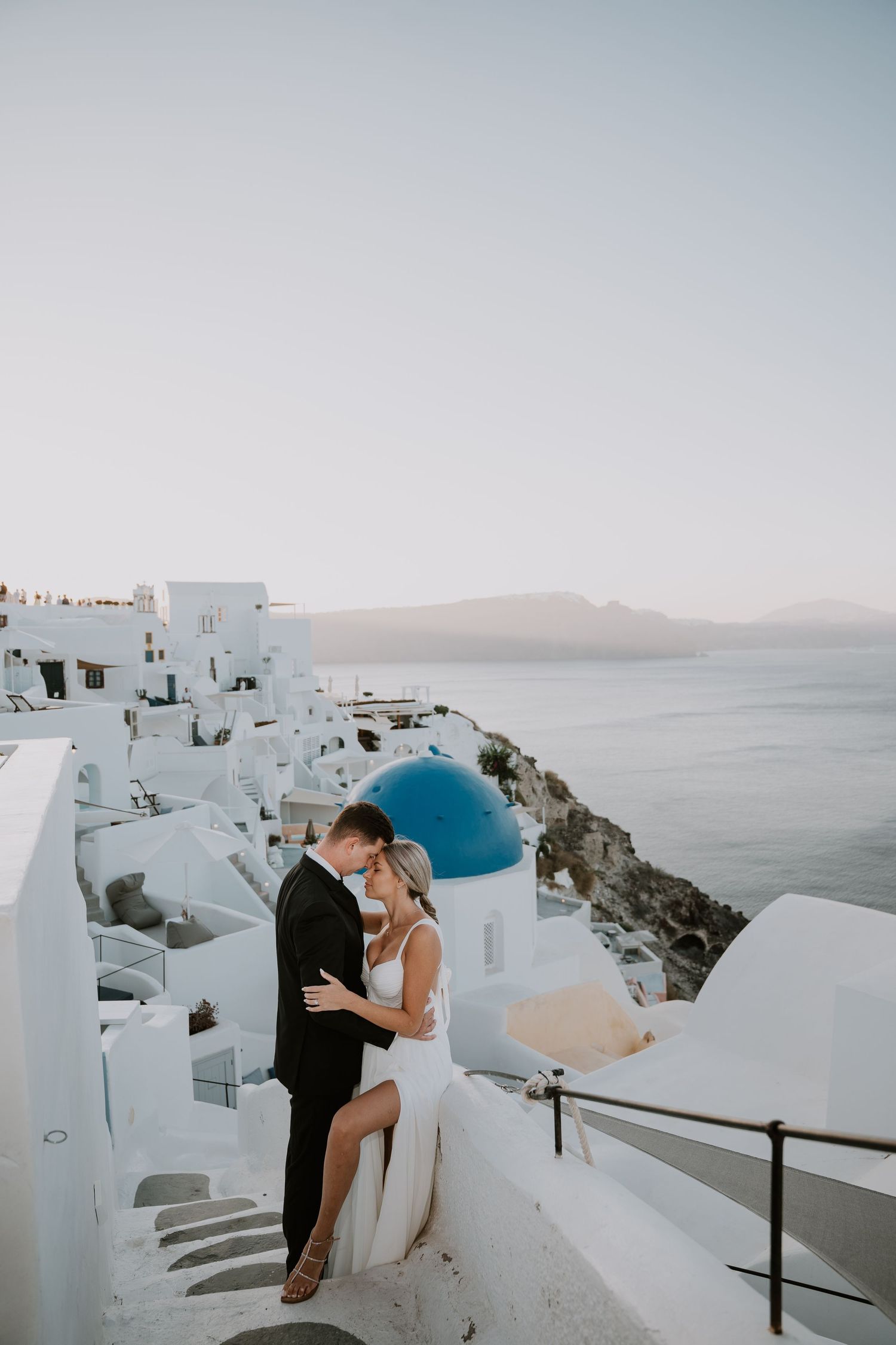 A couple in wedding attire poses on white steps with iconic Santorini blue domes and sea views.