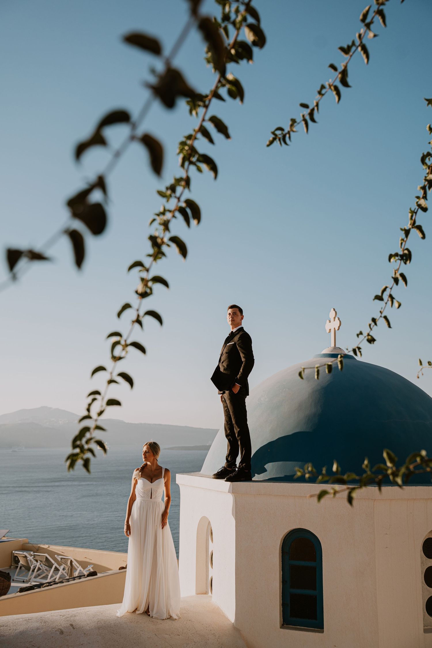 A couple posing by traditional white Greek architecture with blue domes against a clear sky.