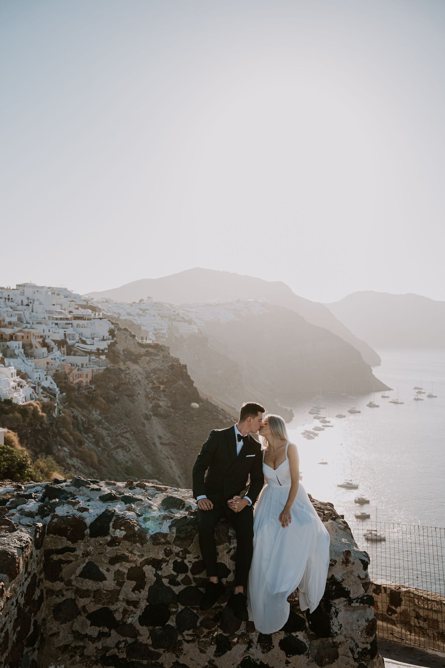 A couple shares an intimate moment on a cliff overlooking the stunning coastline of Santorini at sunset.