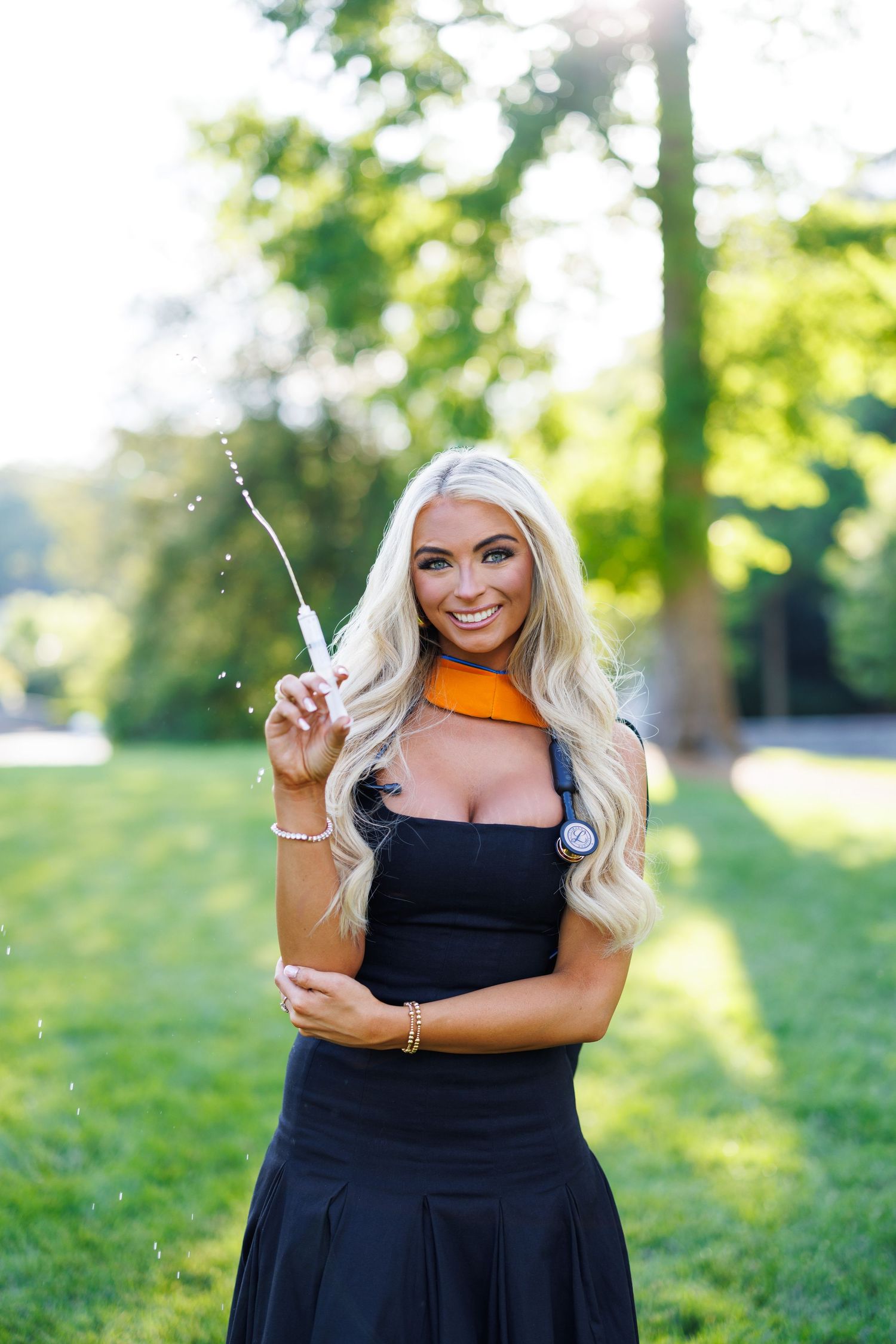 Smiling blonde woman in black dress with orange scarf holds sparkler outdoors in lush green park setting.