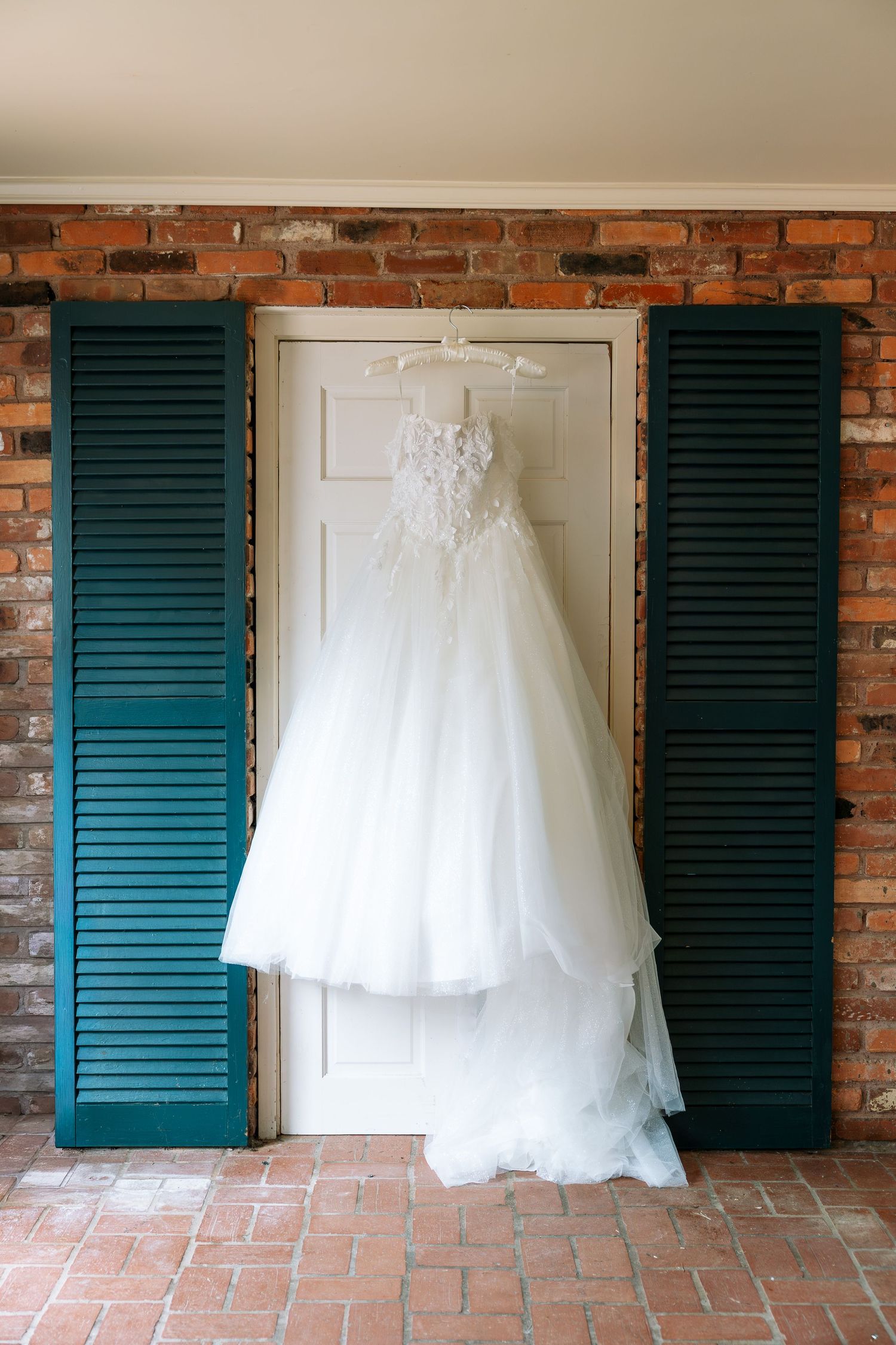 White lace wedding dress hanging on a white door flanked by teal shutters against a rustic brick wall.