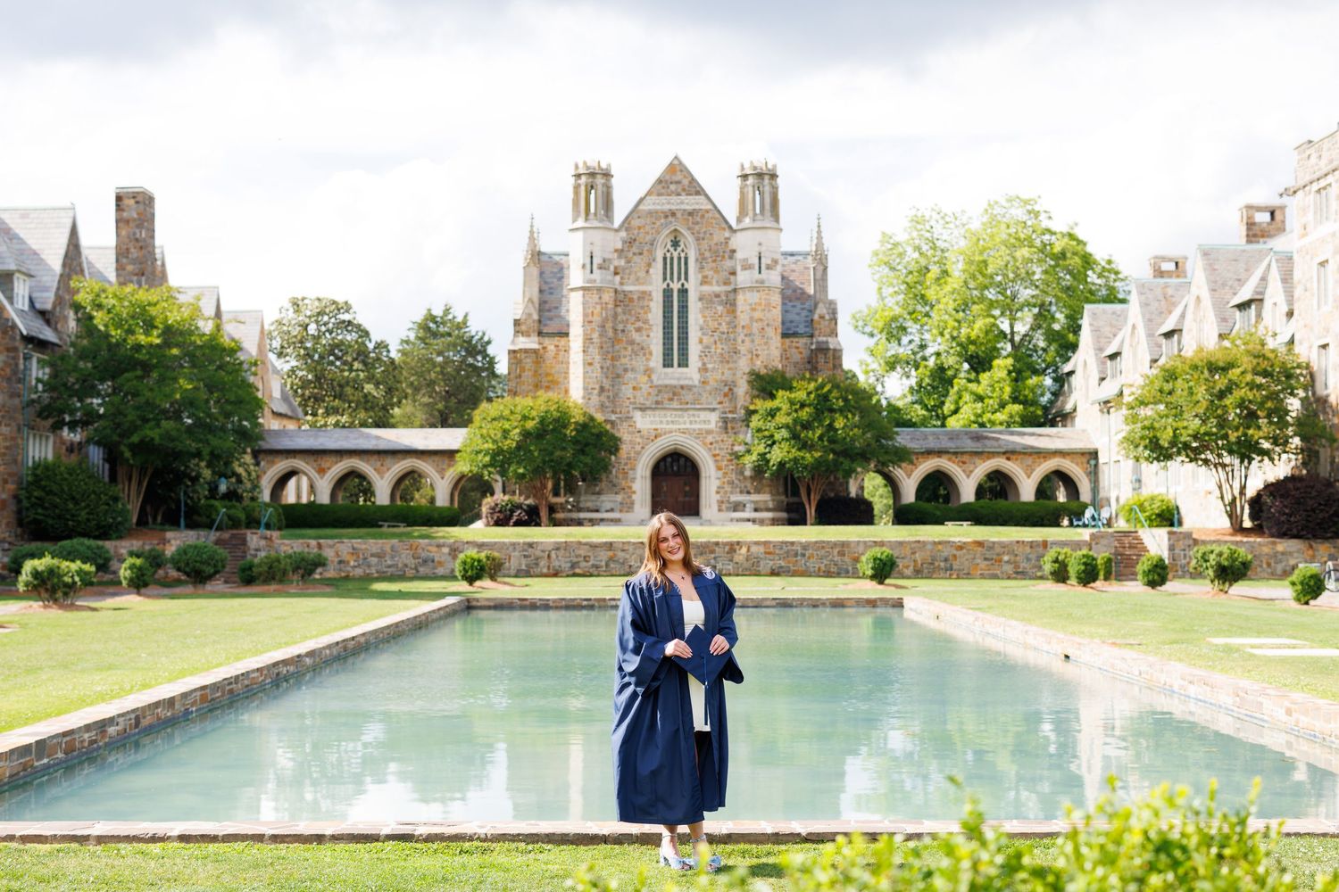 Graduate in cap and gown poses before a Gothic stone chapel with reflecting pool at a university campus.