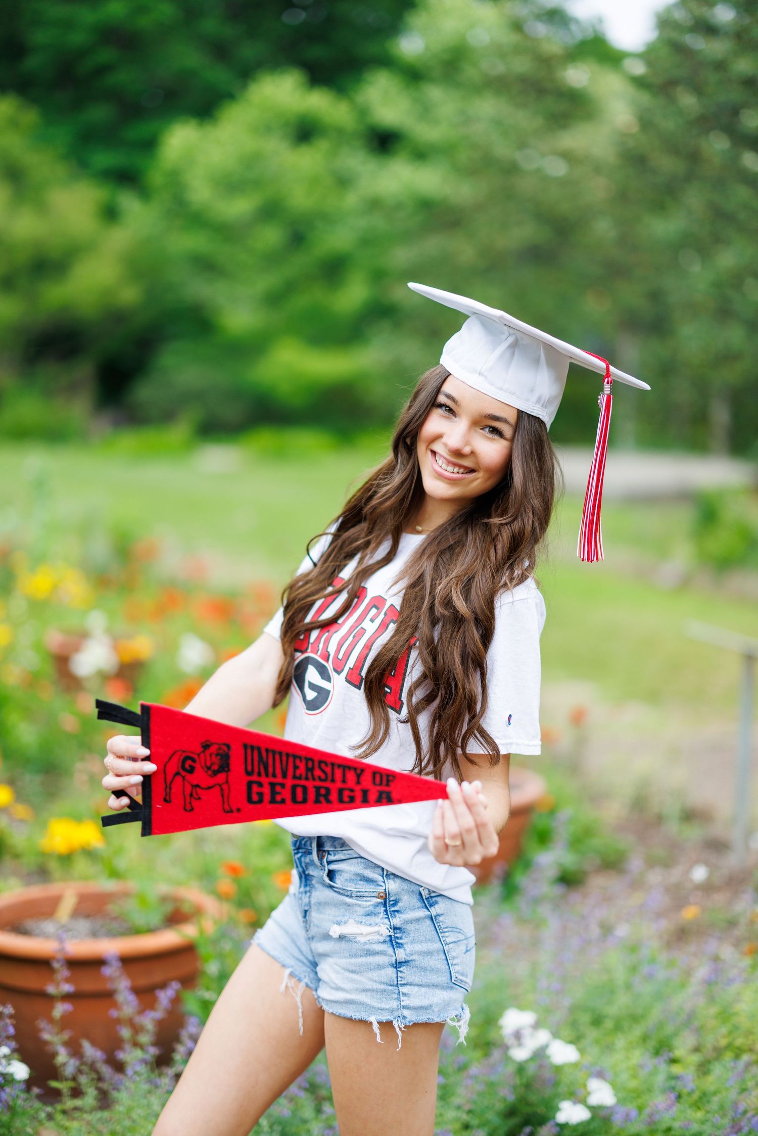 Smiling graduate in white cap holding University of Georgia pennant and wearing UGA shirt with denim shorts outdoors.