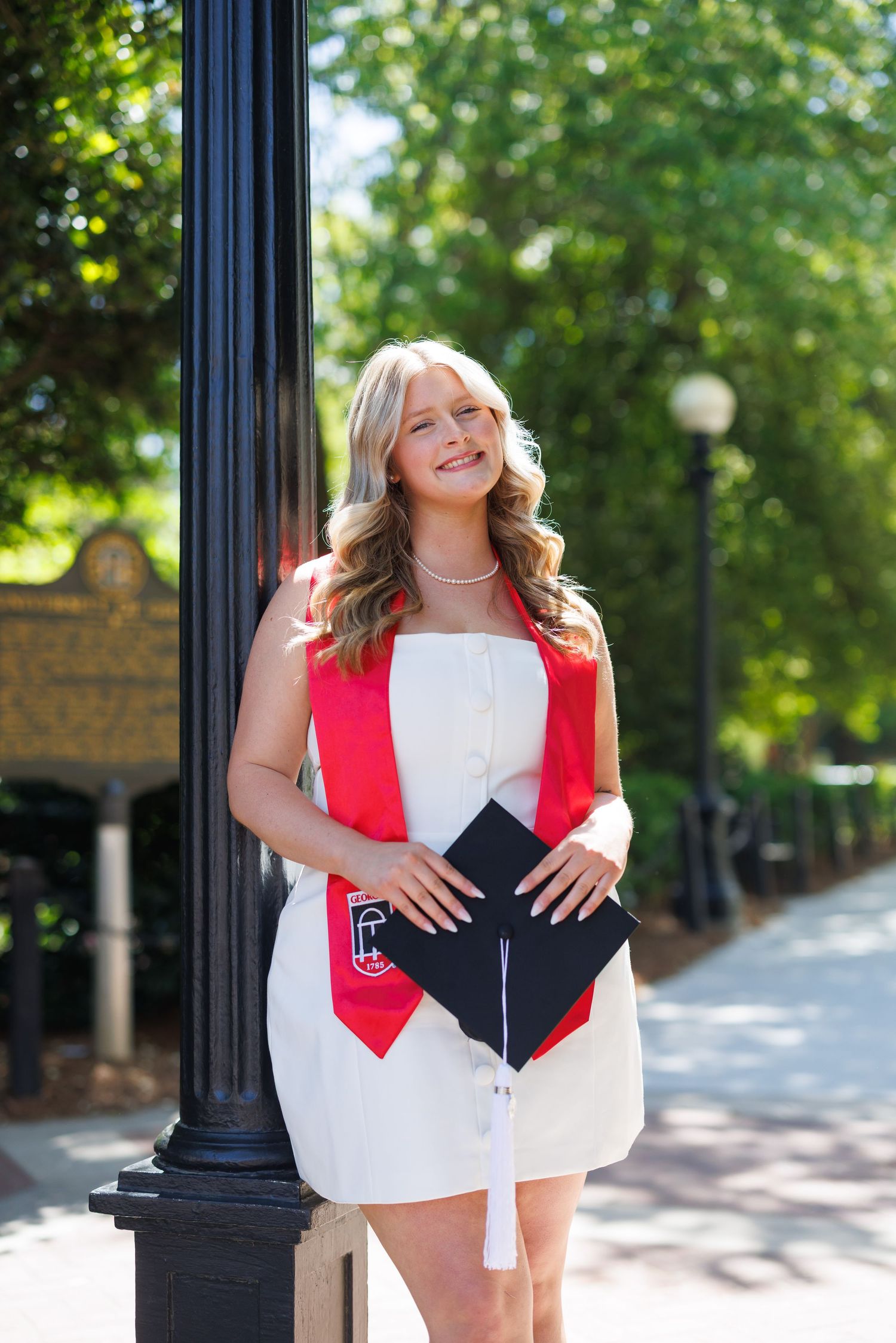 Smiling graduate in white dress with red stole holds cap beside lamppost outdoors.