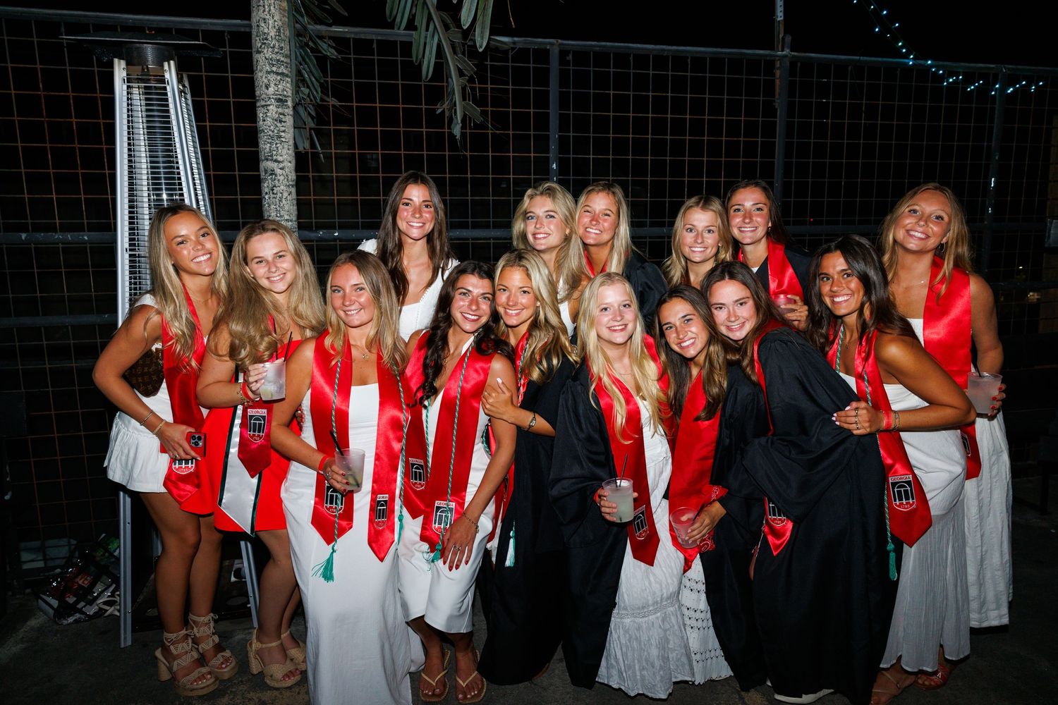 Group of young women in white dresses and red sashes posing together outdoors at a graduation celebration.