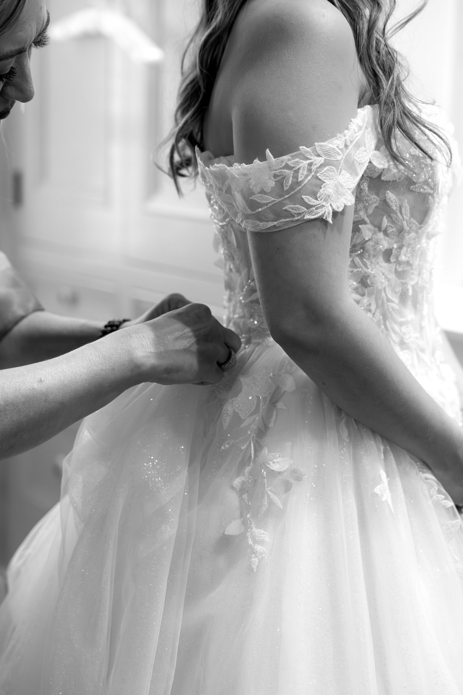 Black and white photo of a bride in a floral lace off-shoulder wedding gown being buttoned up by a helper.