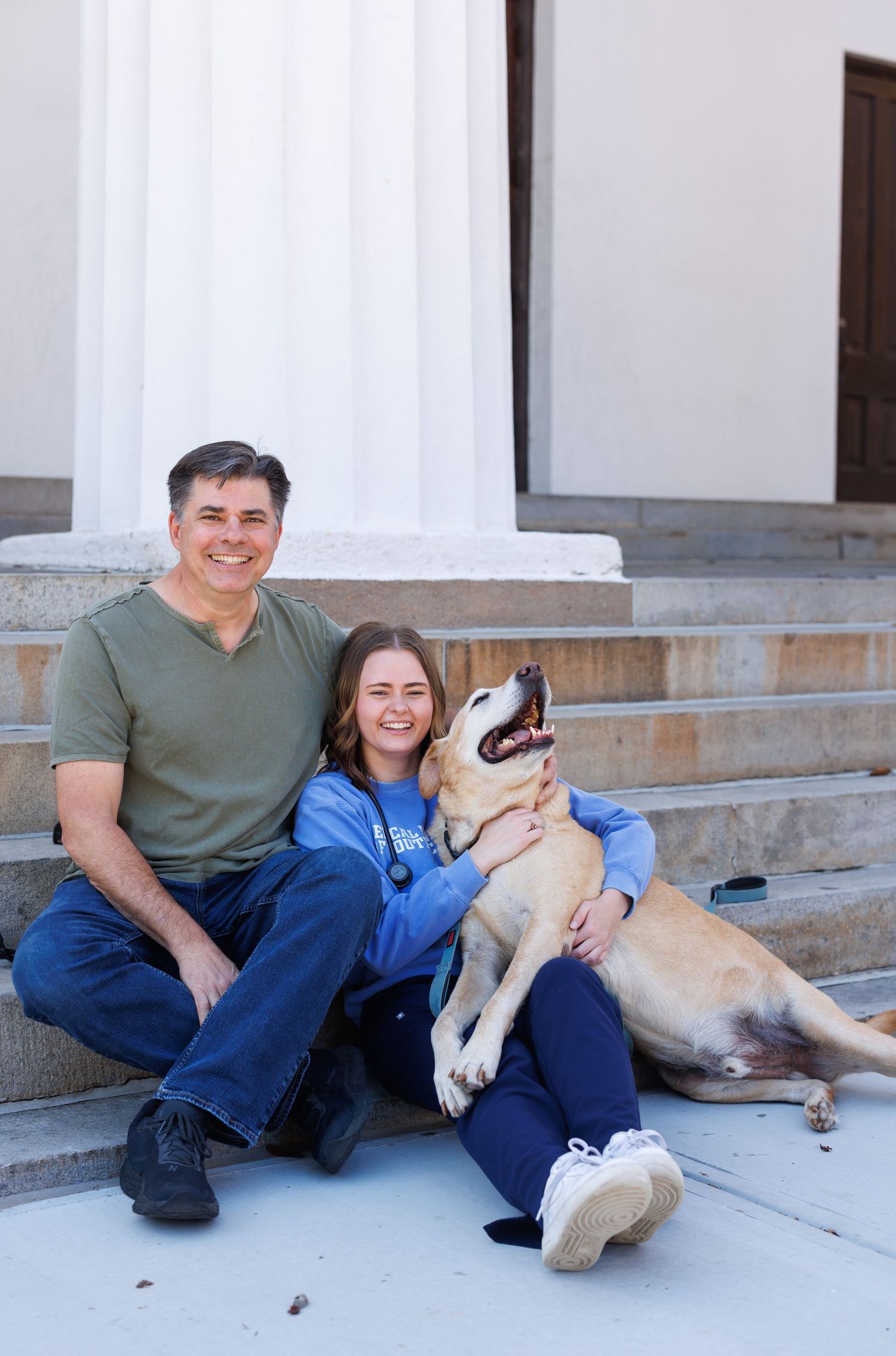 A couple sits with their tan dog on outdoor stone steps near white columns.