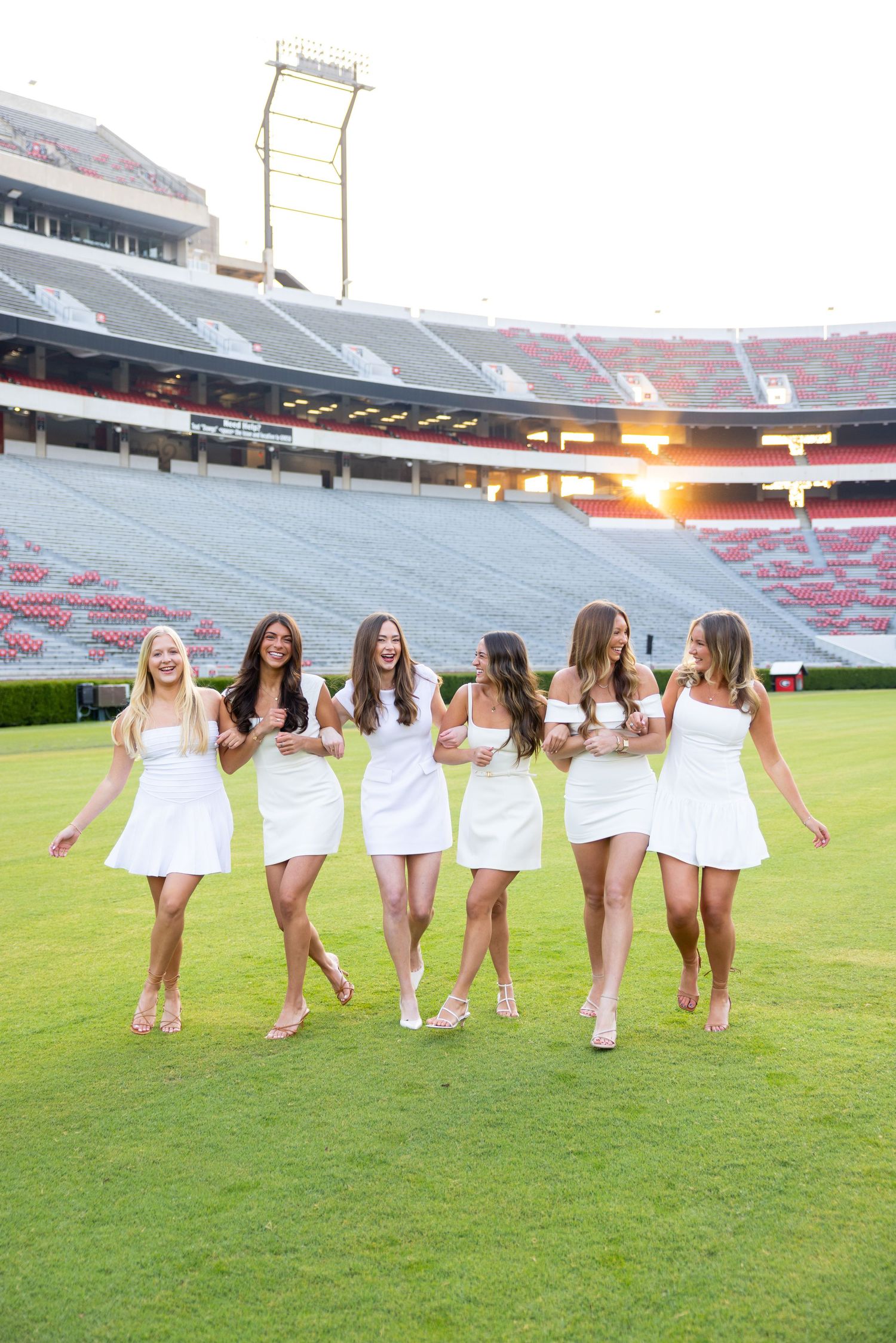 Six women in white dresses walking on a football field at sunset in a large stadium.