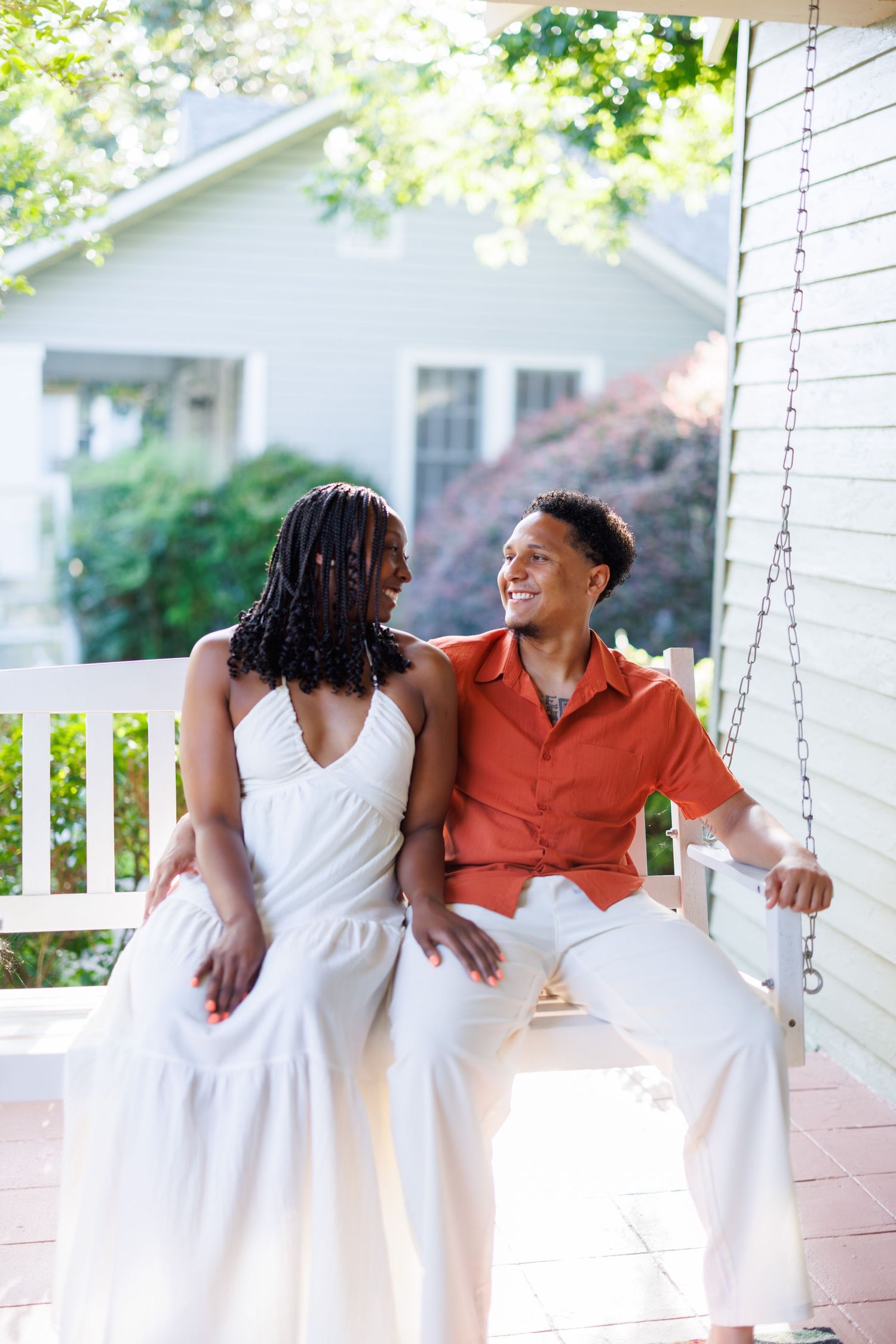 A happy couple sits on a porch swing, woman in white dress and man in orange shirt, sharing a warm smile.