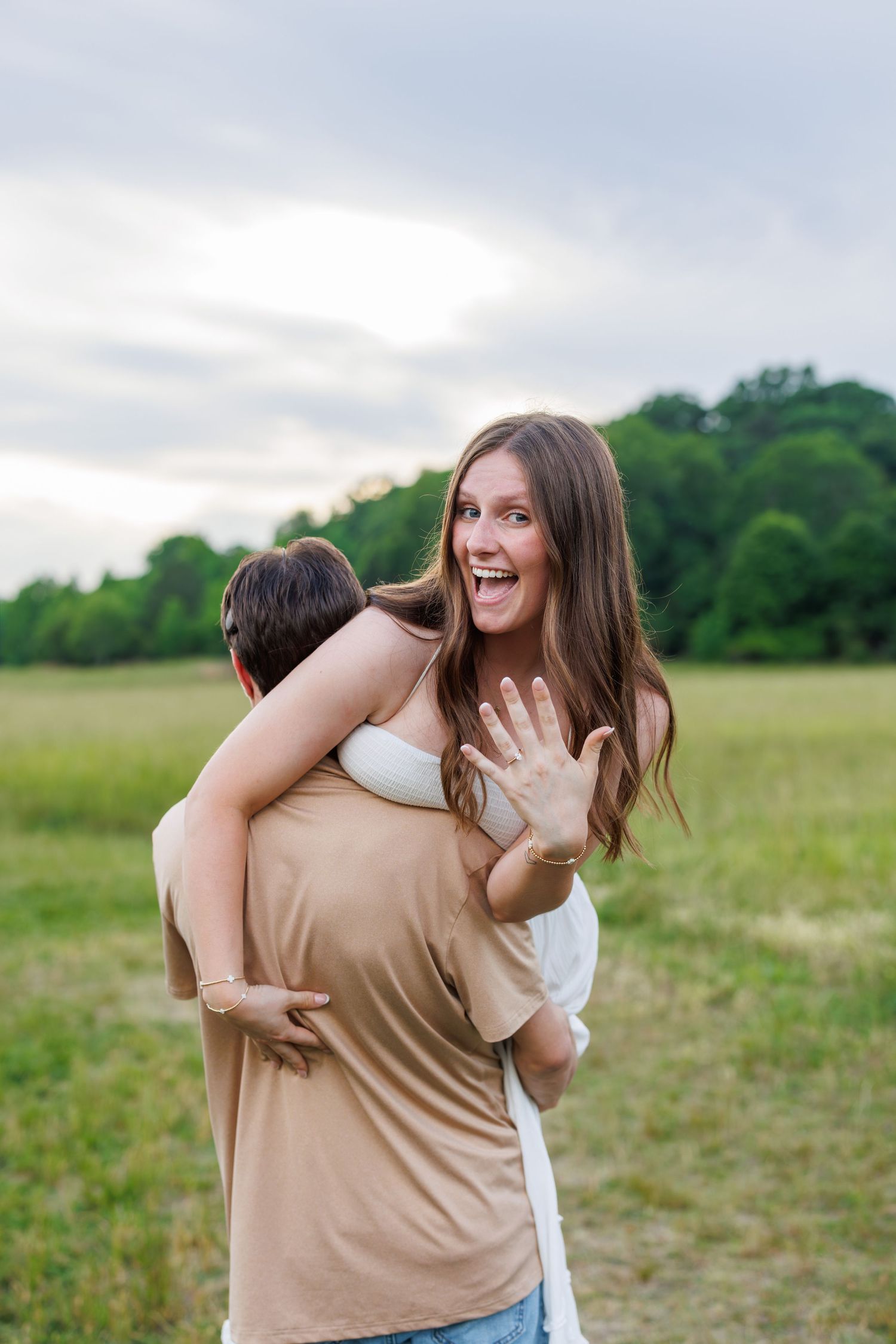 Excited woman shows off engagement ring while being hugged by her partner in a green field.