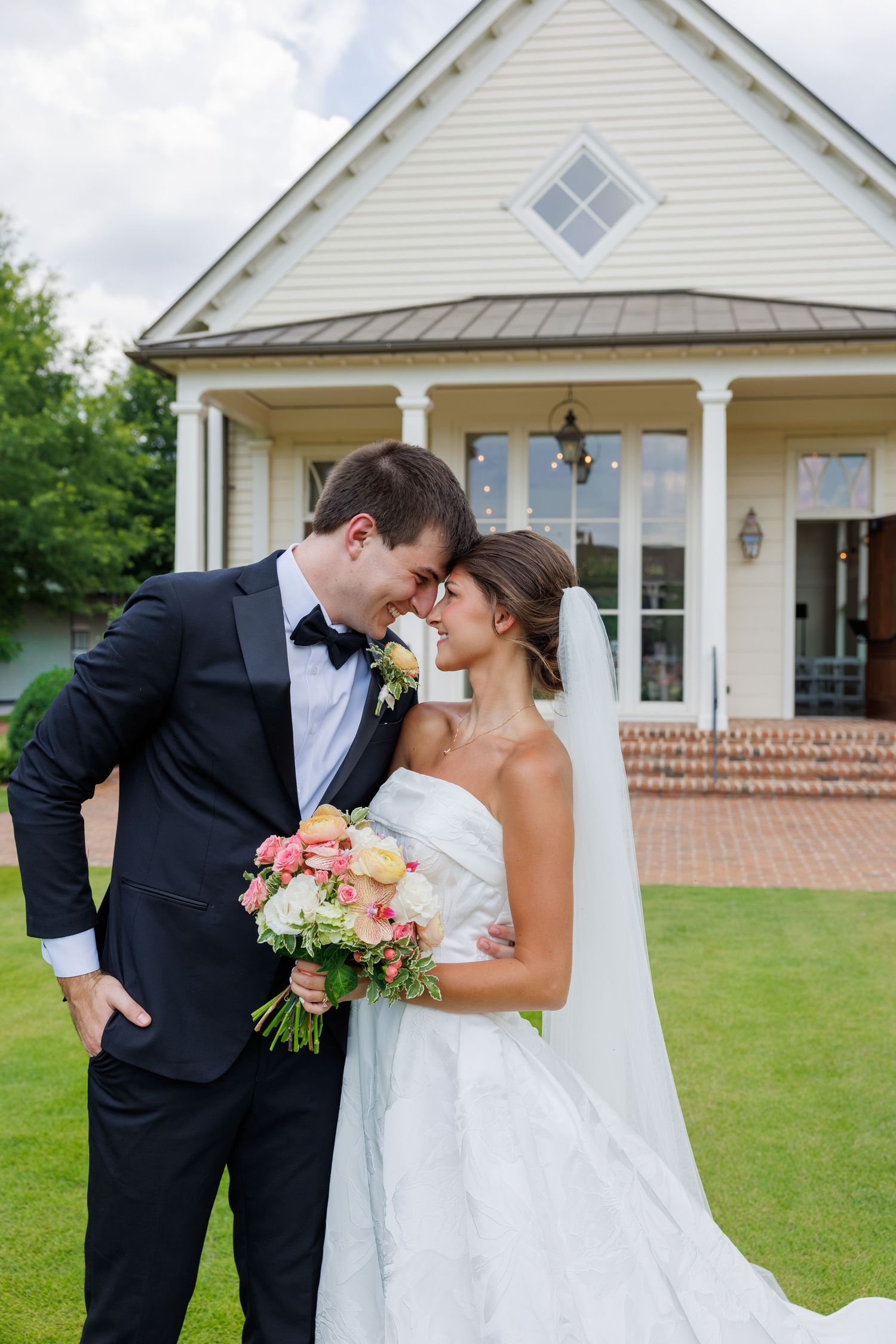 Bride and groom share a romantic moment outside a white chapel, bride holding a pink and white bouquet in a strapless gown.