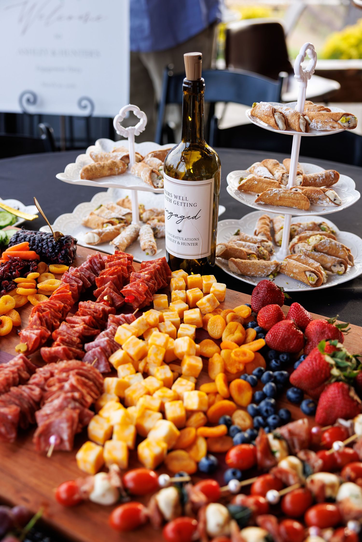 Elegant charcuterie board with cheese, fruits, meats, and a wine bottle at a catered outdoor event.