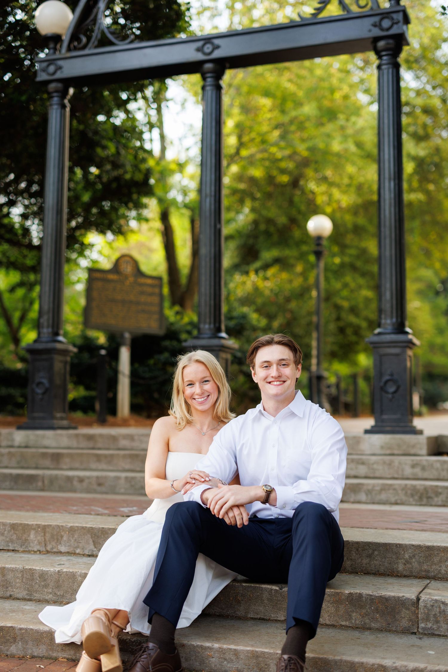 A couple relaxes on stone steps near ornate black lampposts in an urban park setting.