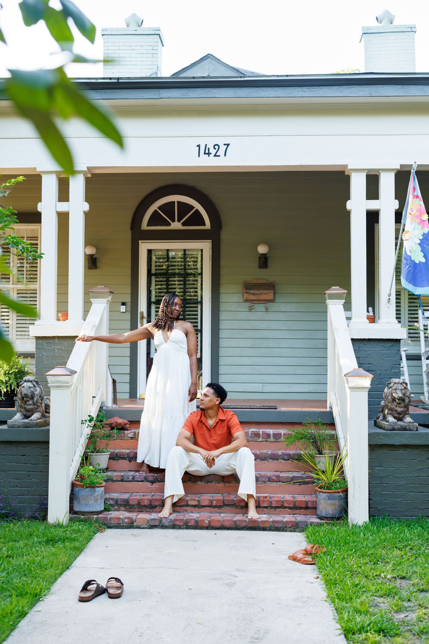 Couple posing on brick steps of a charming gray house numbered 1427, woman in white dress, man in orange shirt.