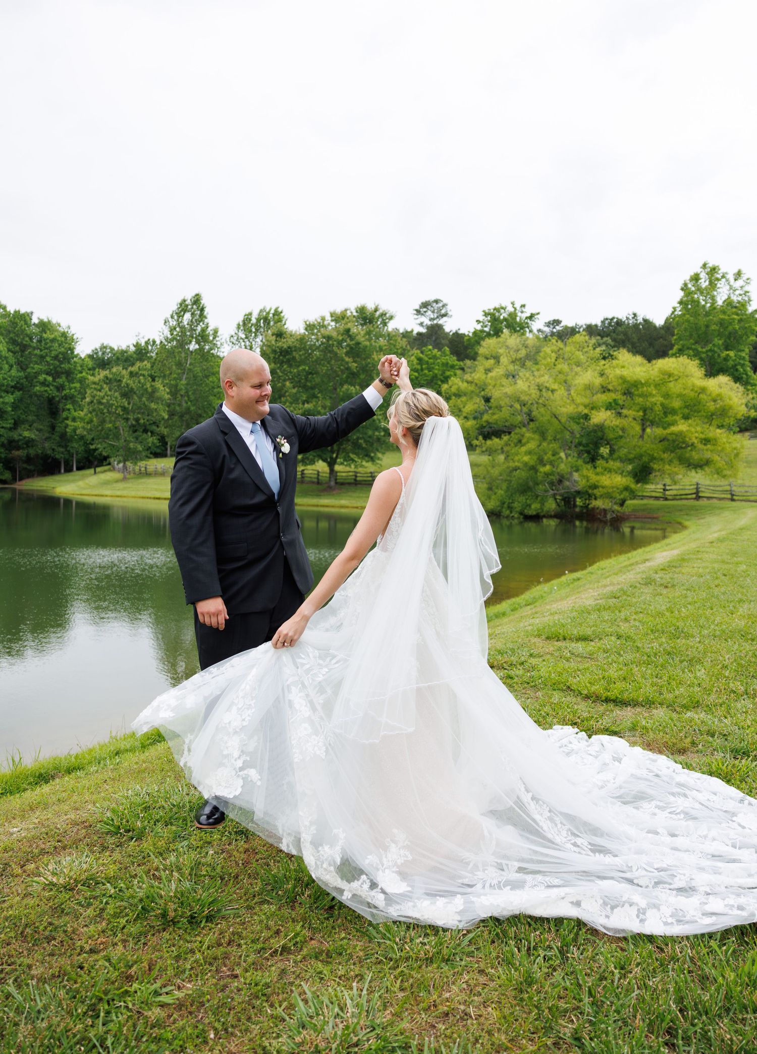 Wedding couple walking along a grassy hillside by a lake with a flowing white wedding dress train.