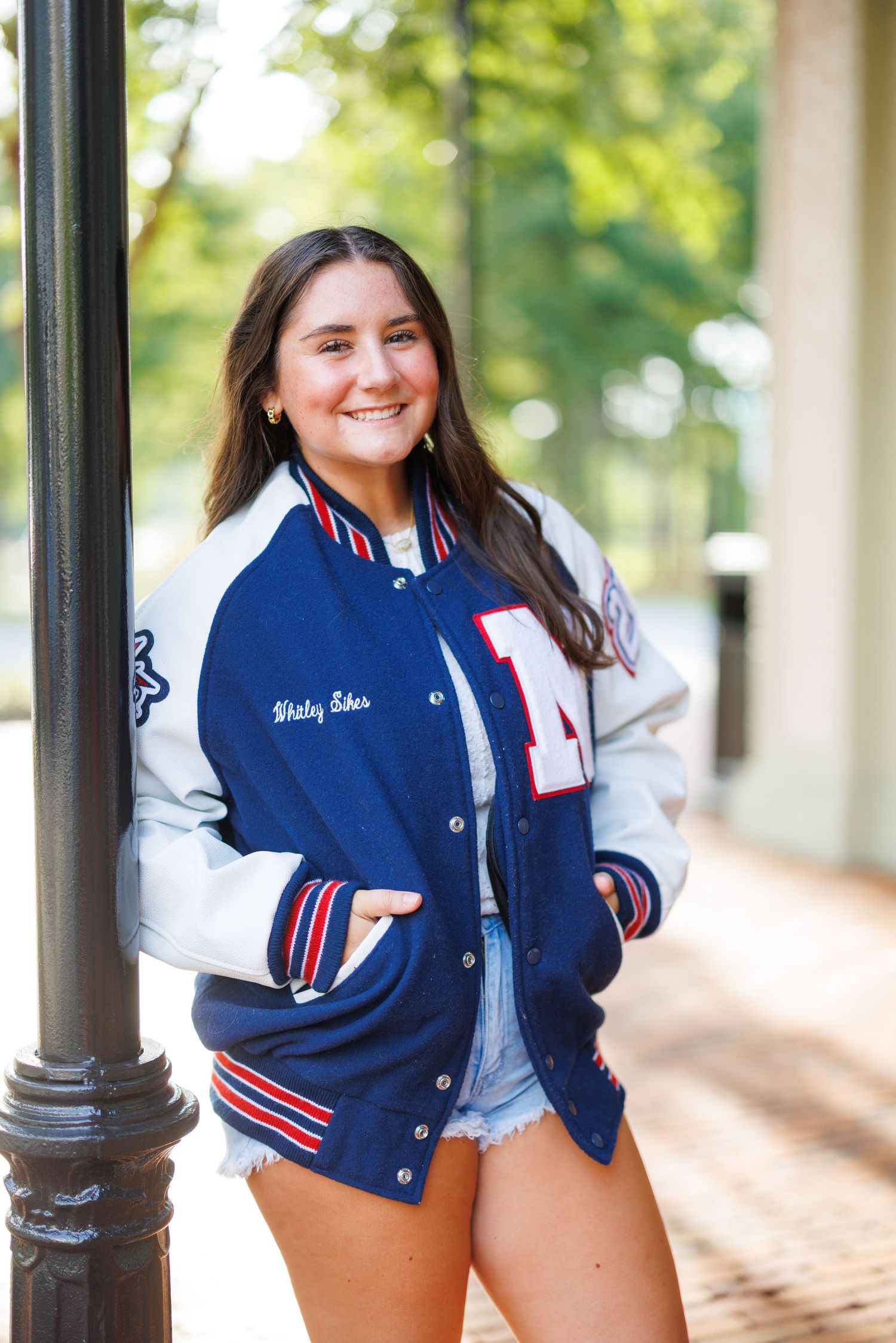A person wearing a blue and white varsity letterman jacket poses outdoors near black lamp posts.