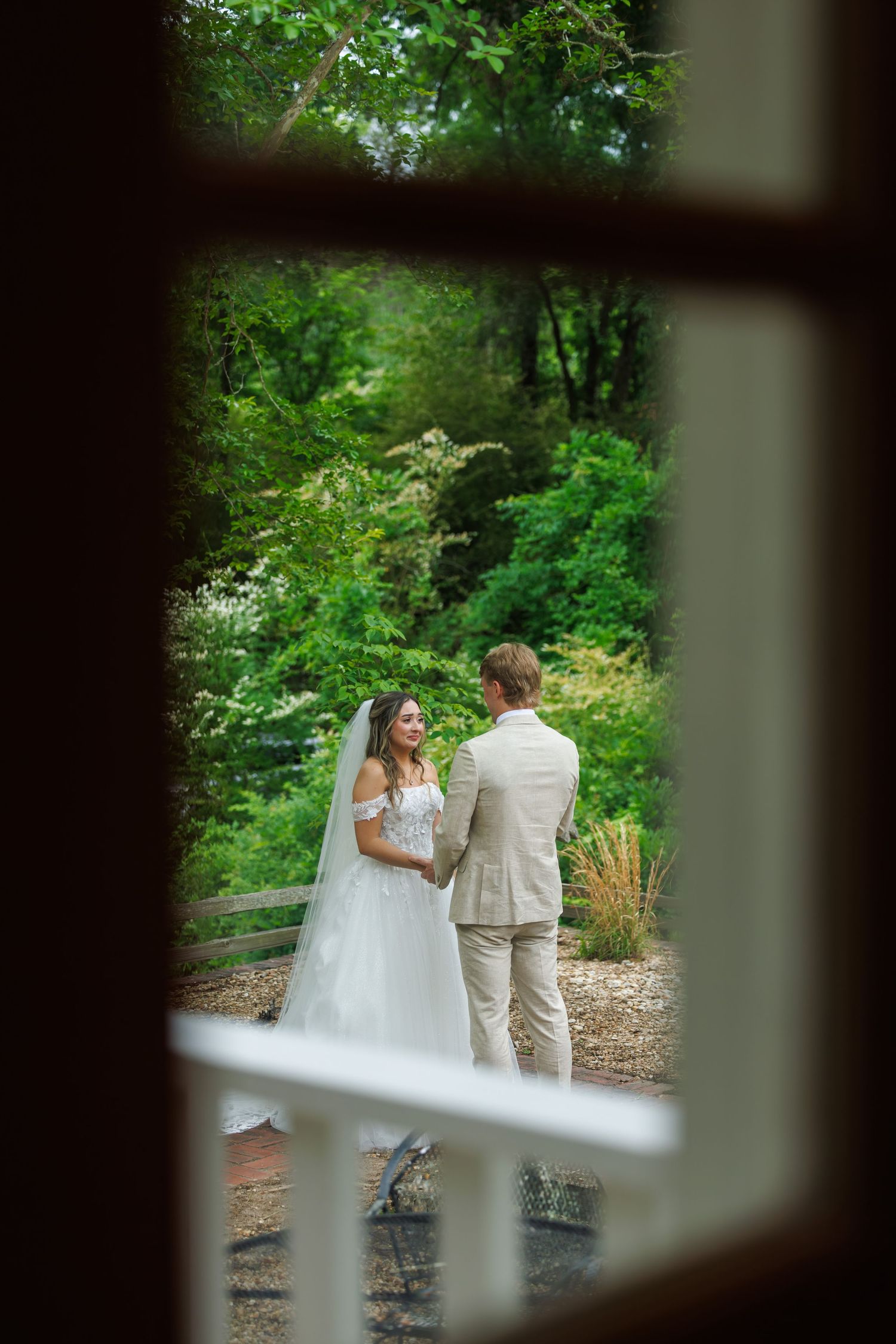 Bride and groom sharing a romantic first look moment in a lush green garden, viewed through a window.