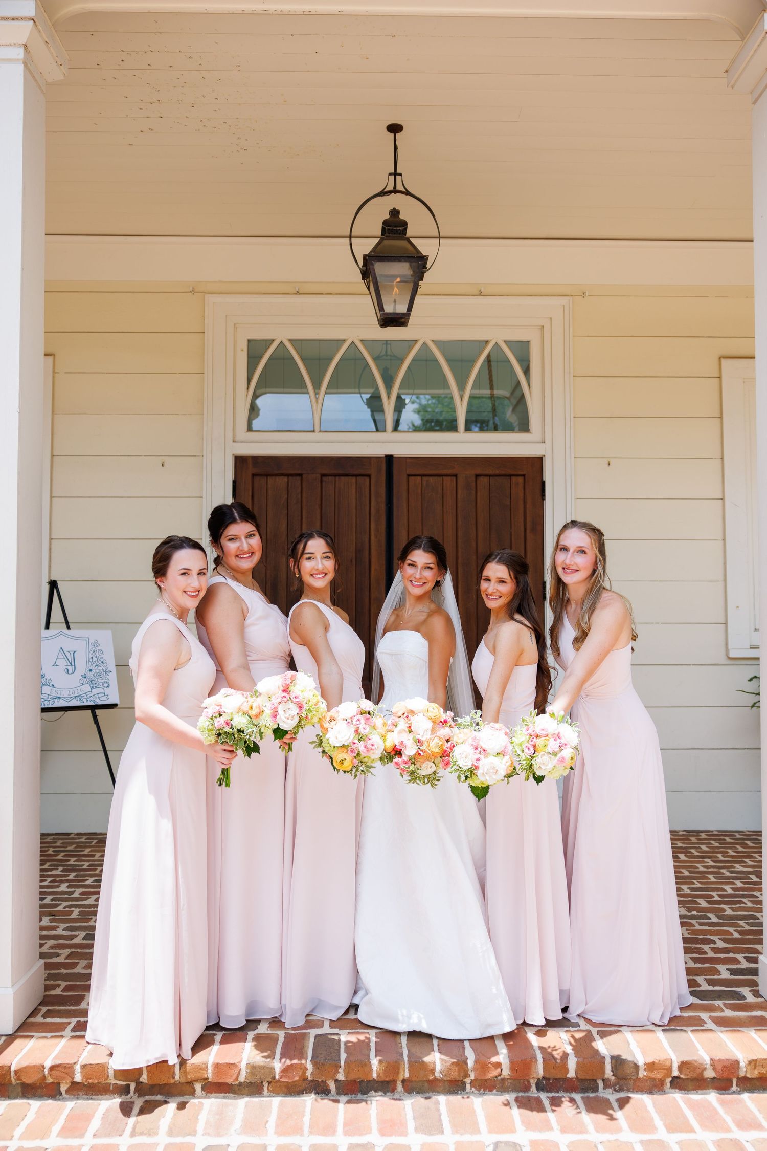 Bride in white gown with five bridesmaids in blush pink dresses holding floral bouquets on chapel steps.