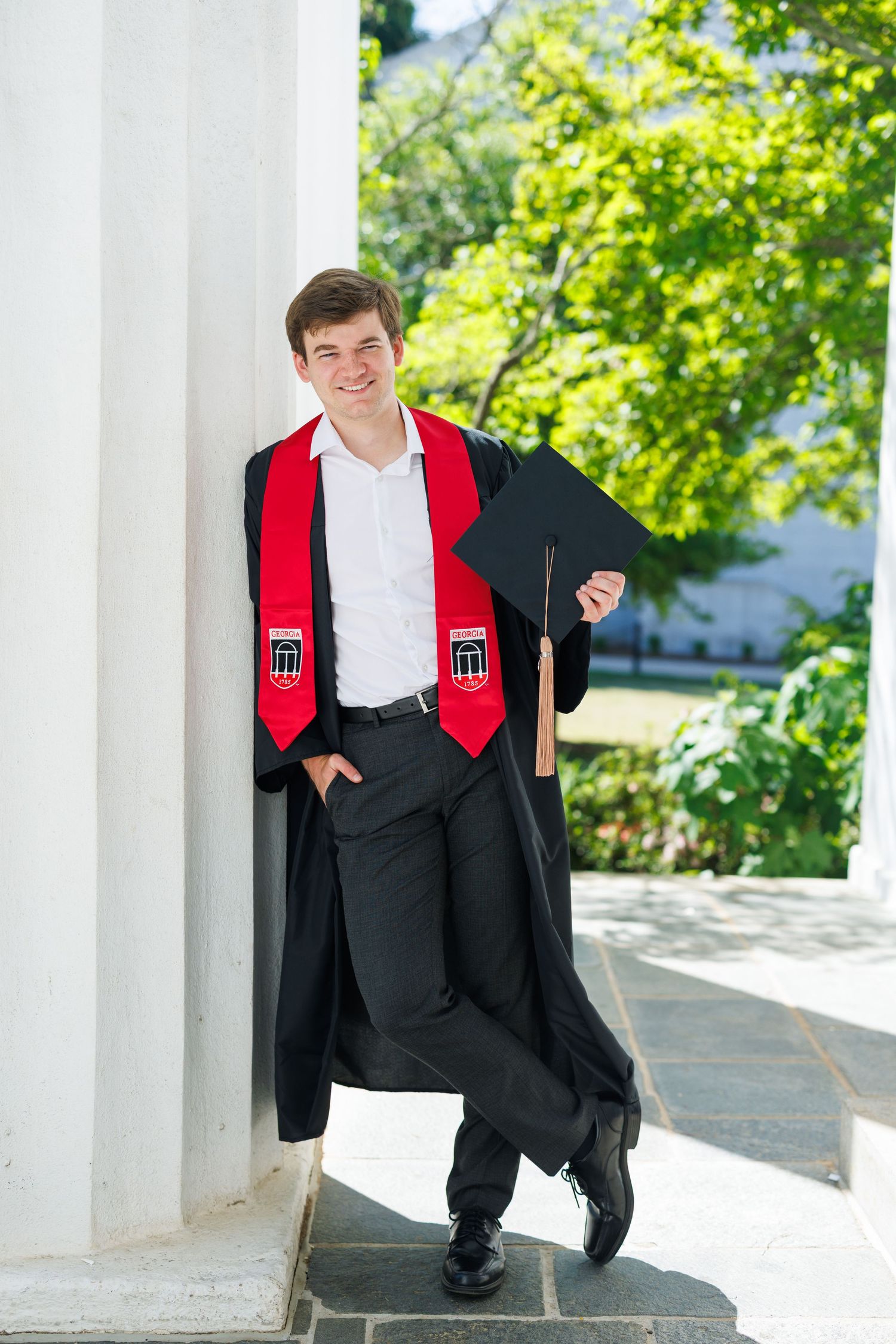 Young man in black graduation gown and red stole holding cap, smiling while leaning against white column outdoors.