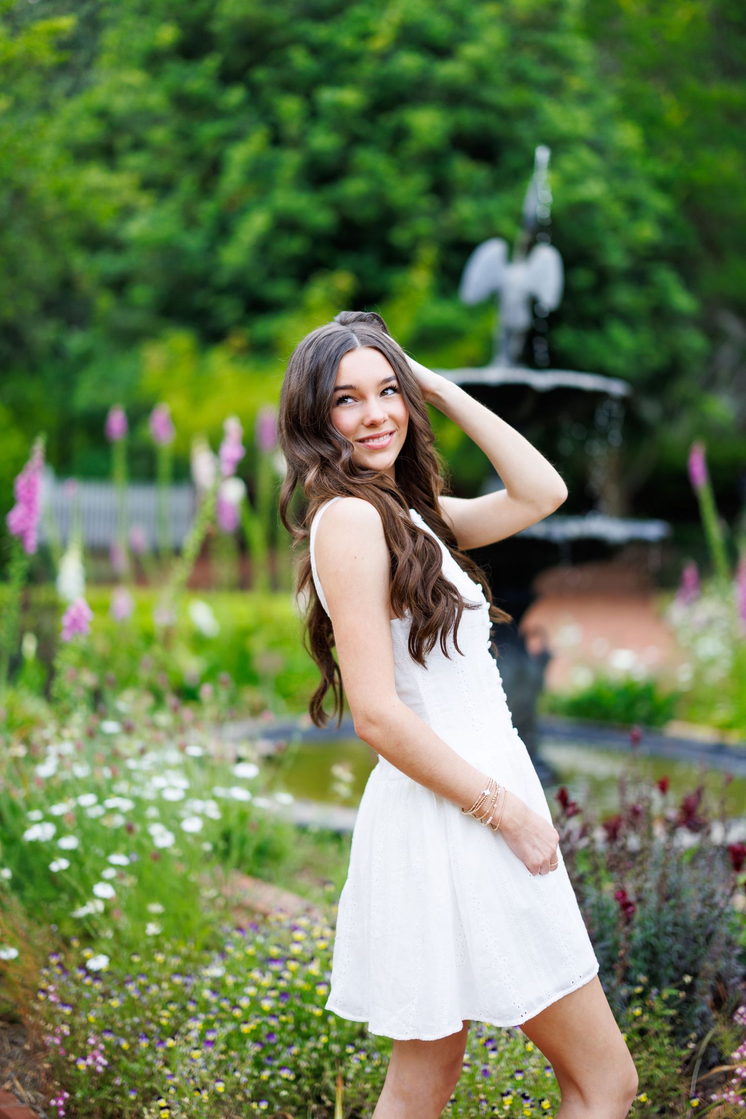 Young woman in white dress posing in a lush garden with a decorative fountain and colorful flowers in the background.