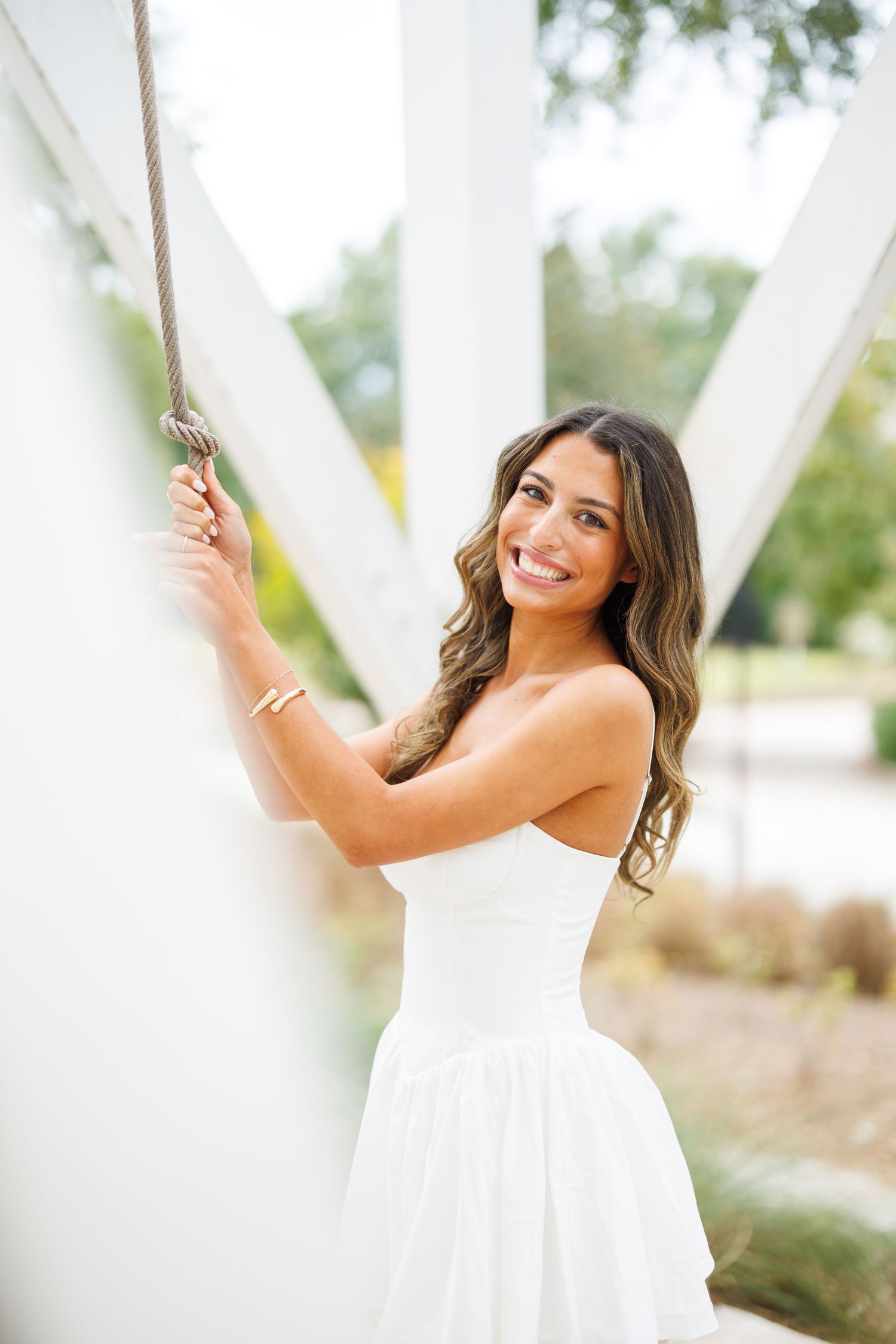 Graduate in white dress poses between white columns during outdoor photo session.