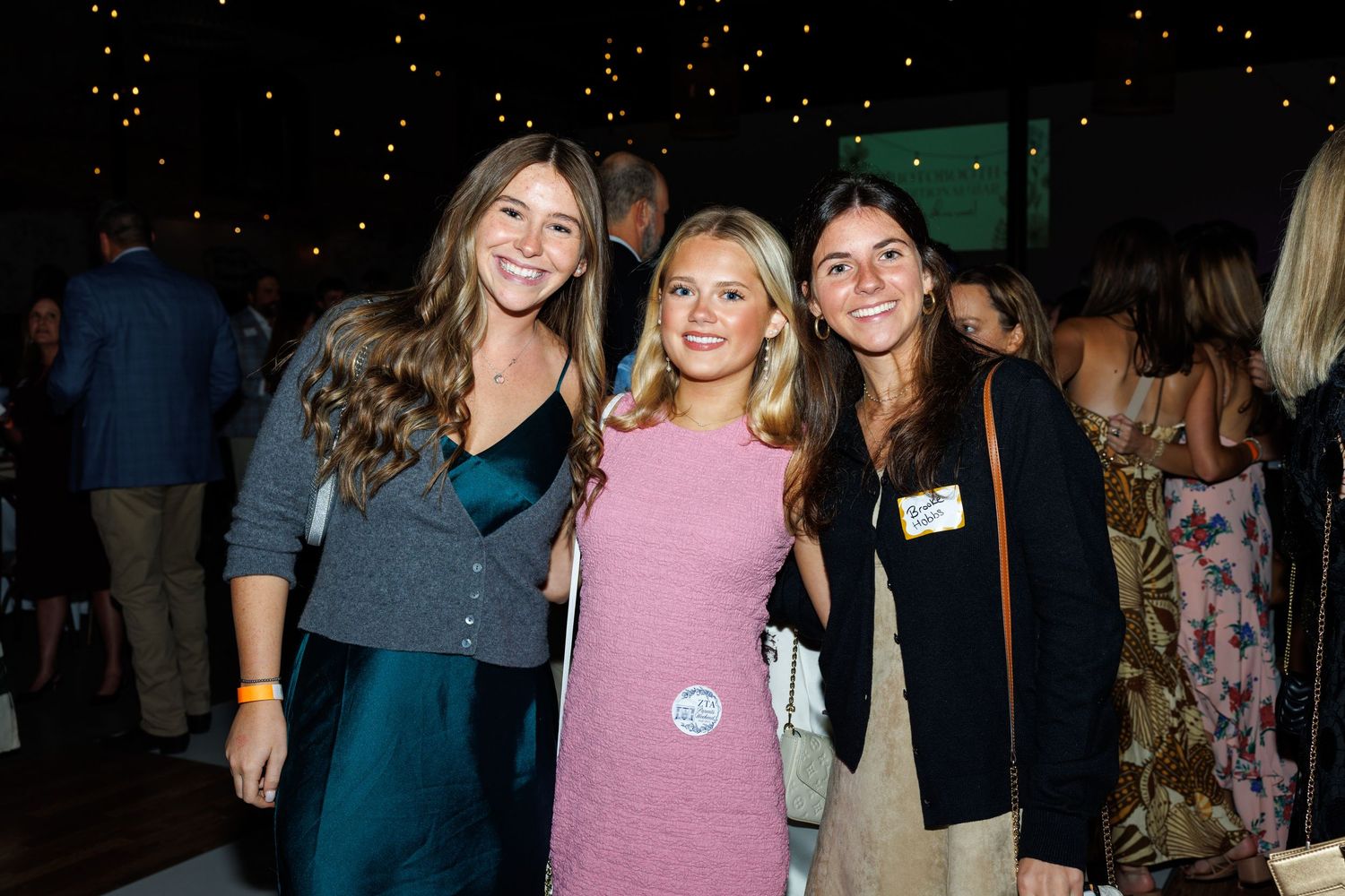 Three smiling young women pose together at an elegant indoor evening event with string lights in the background.