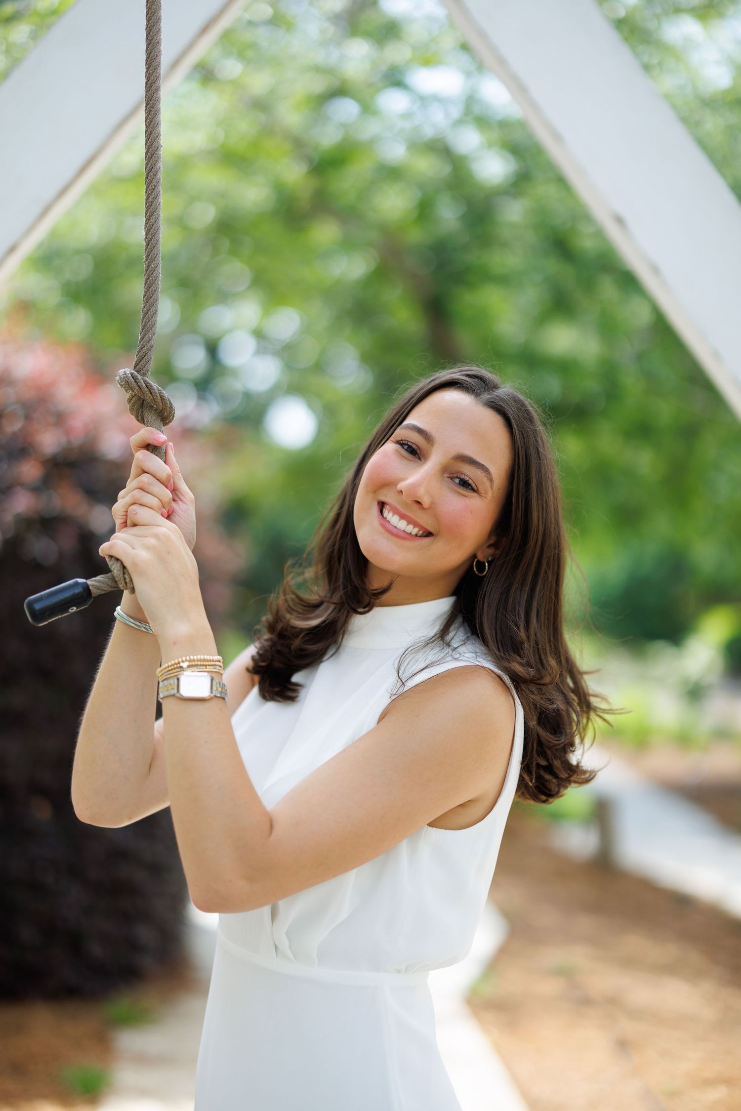 A person in a white sleeveless top smiles while posing on a rope swing outdoors in a natural setting.