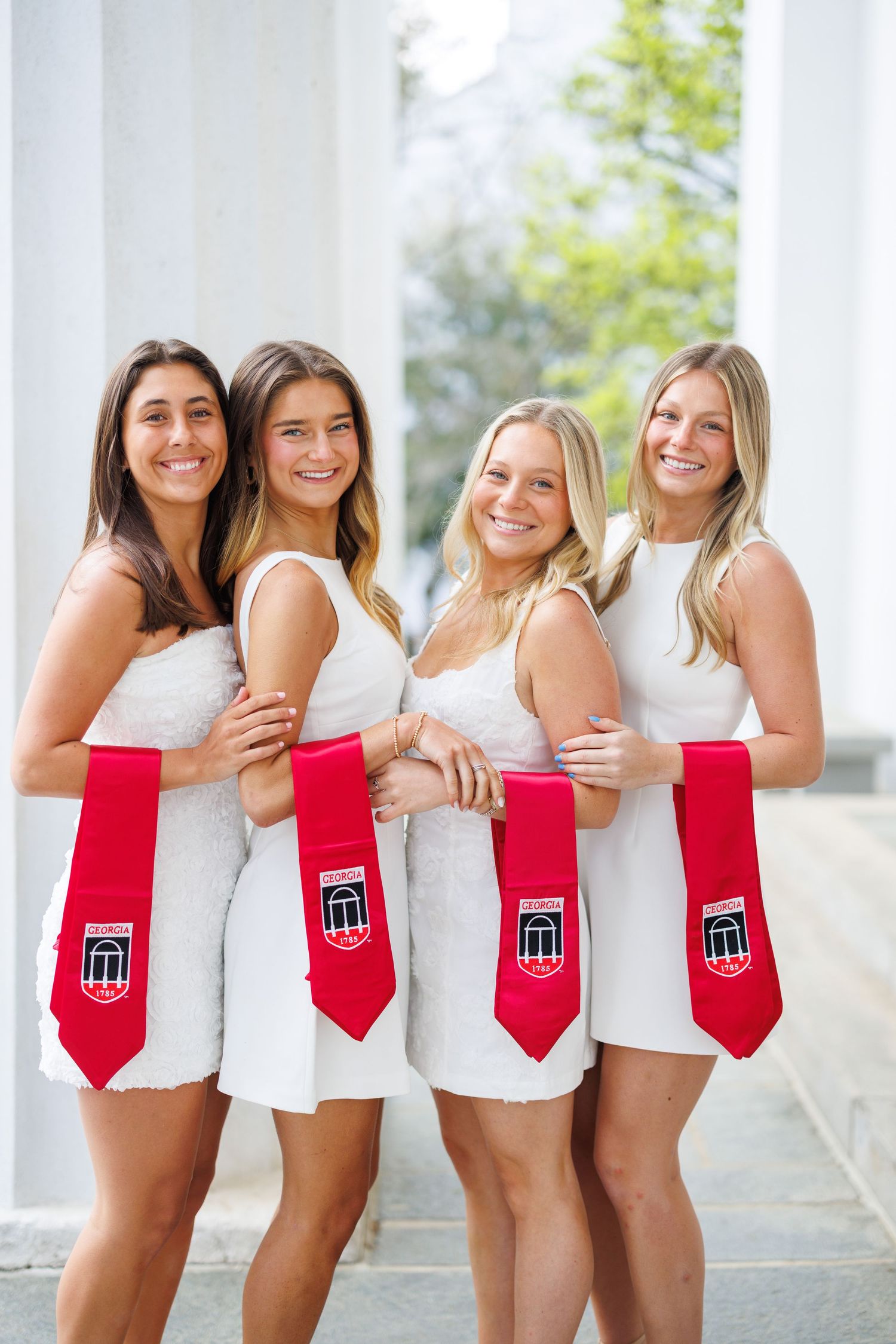 A group of people in white dresses and red sashes pose together for graduation photos.
