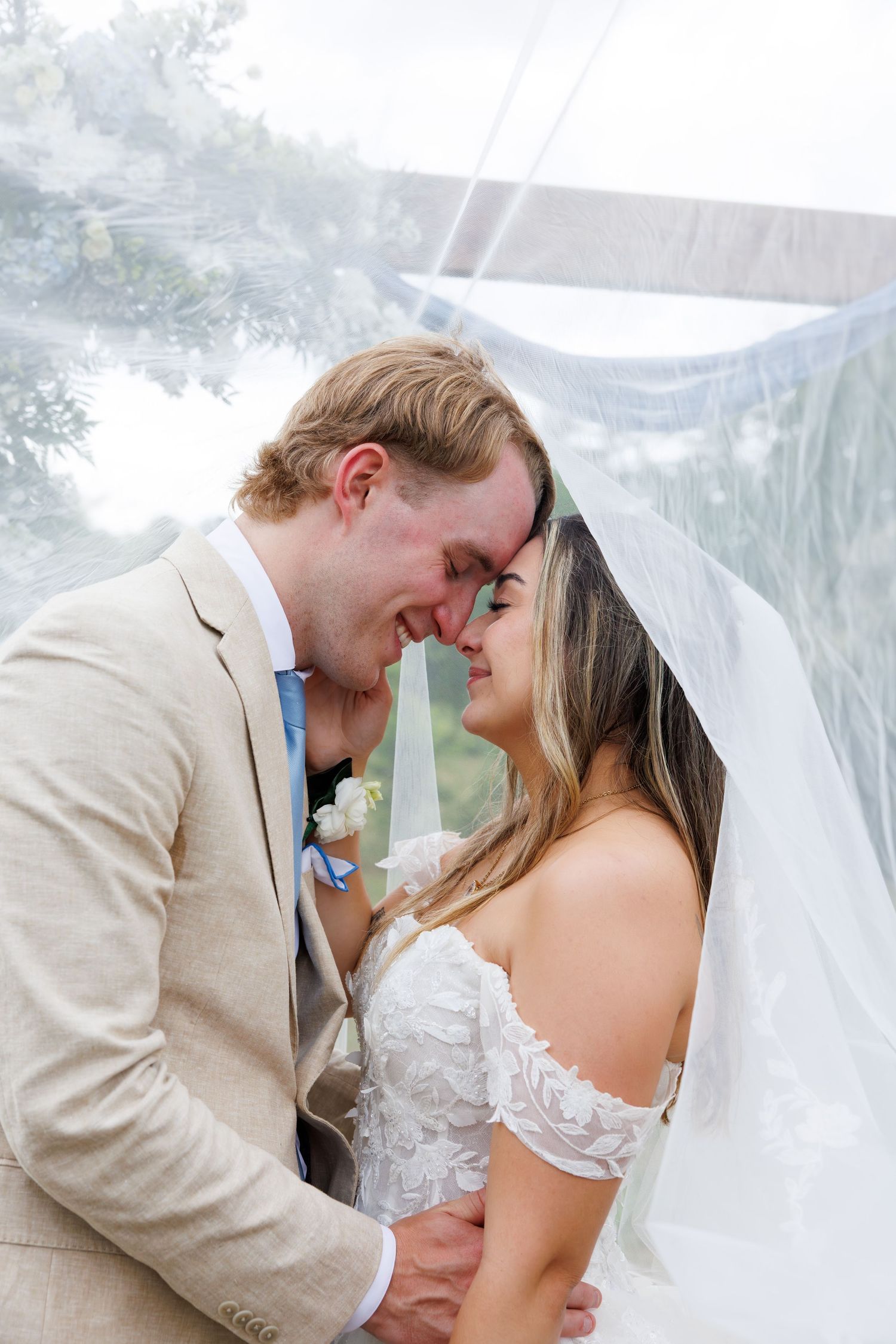Bride and groom sharing an intimate nose-to-nose moment under a flowing wedding veil outdoors.