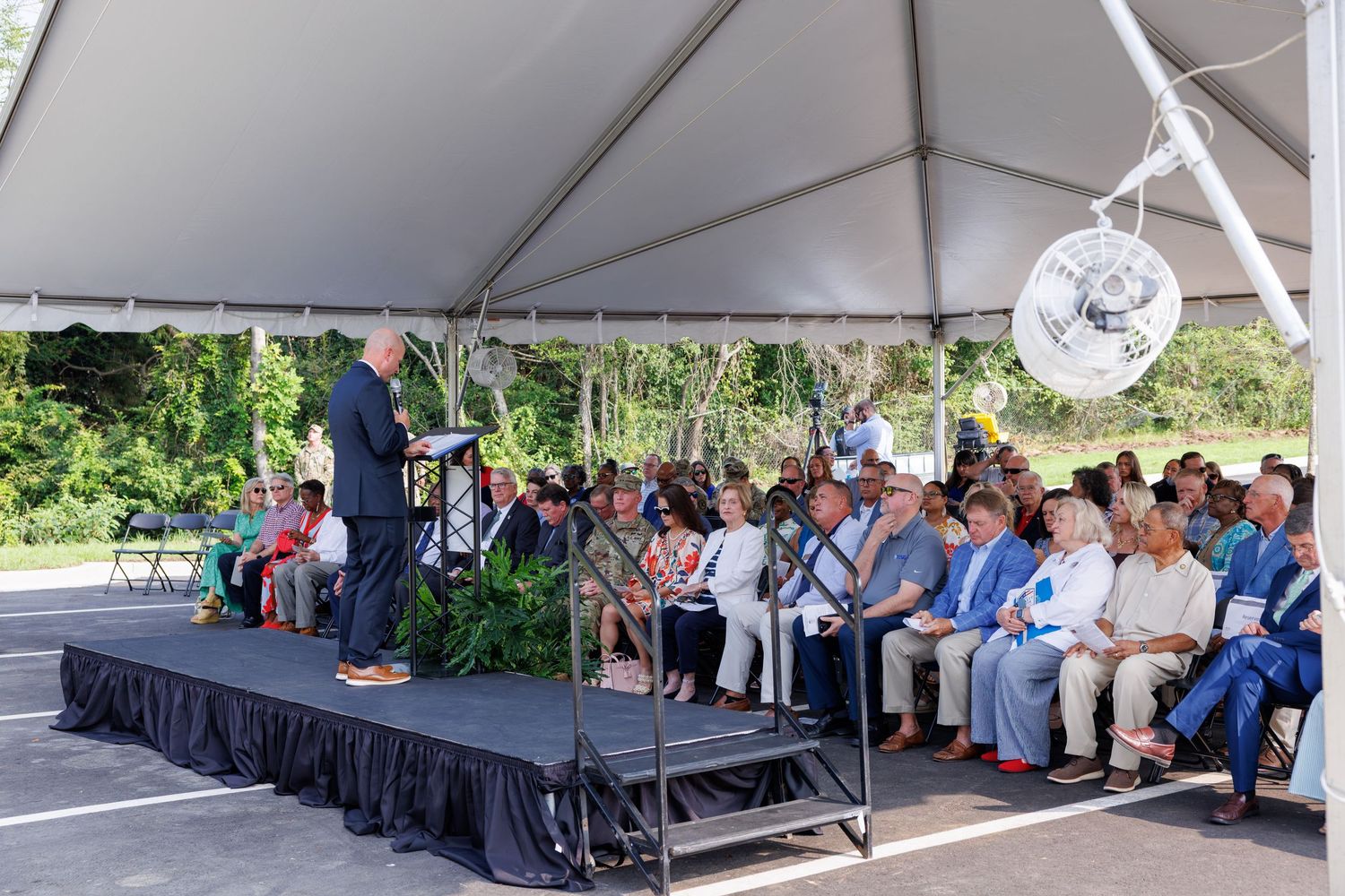 Speaker addresses a large crowd seated under a white tent at an outdoor ceremony event.