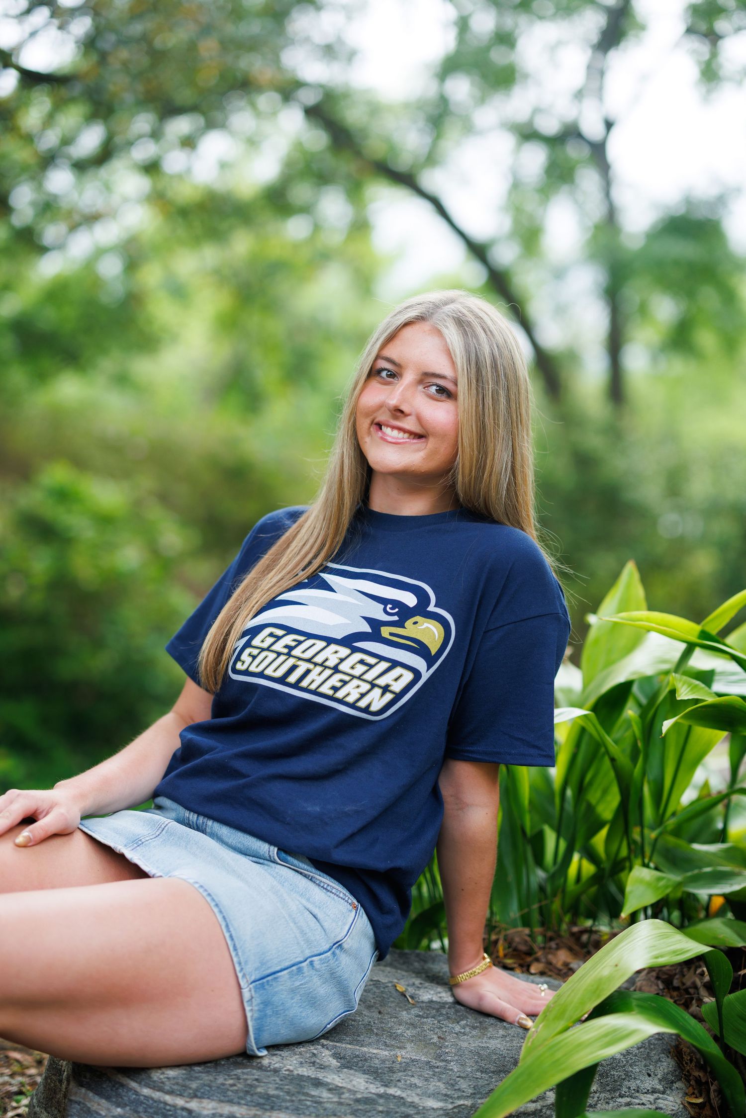 Smiling blonde woman sitting outdoors wearing a navy Georgia Southern Eagles t-shirt and denim shorts surrounded by green foliage.