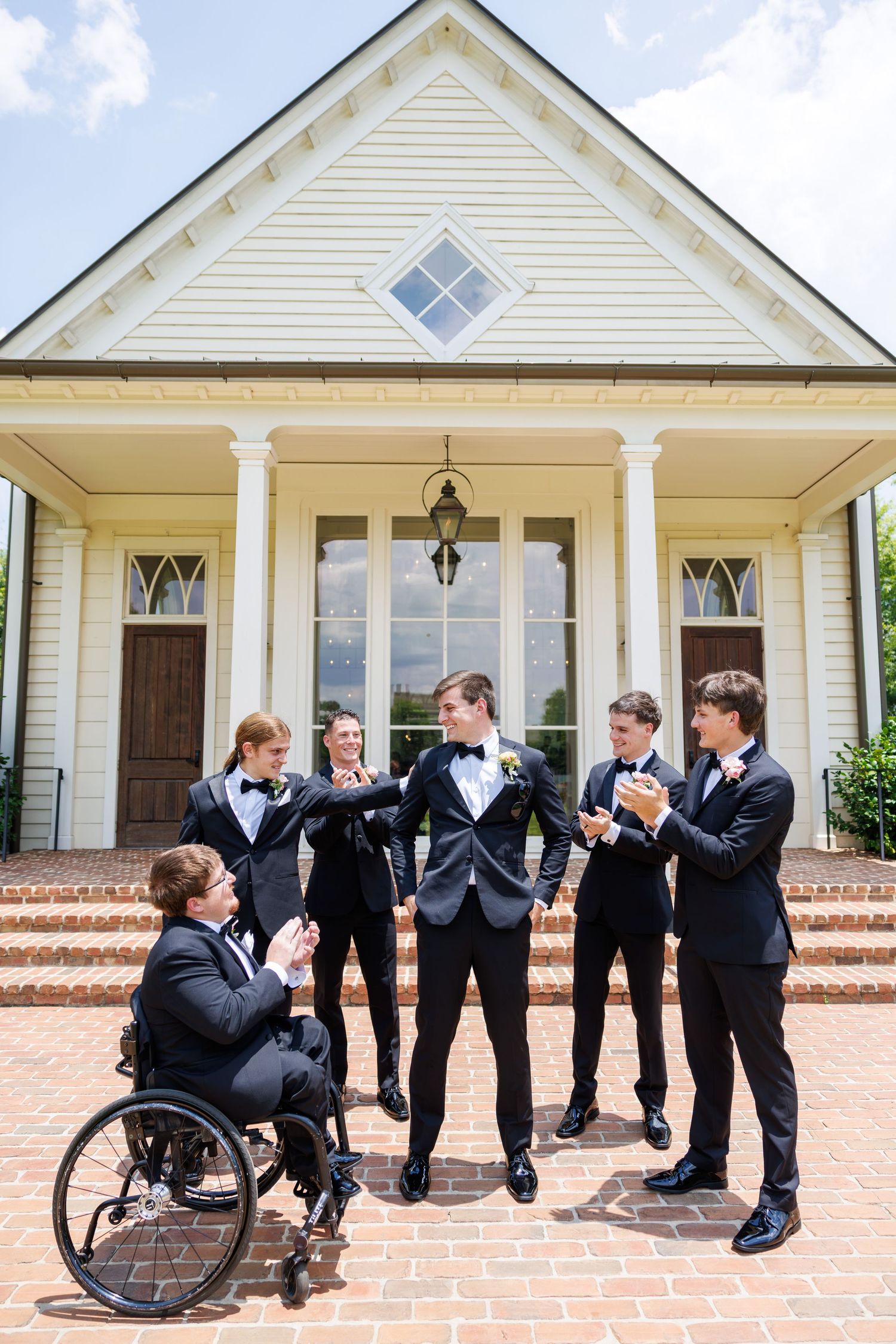 Groom and groomsmen in black tuxedos pose outside a white chapel, including one groomsman in a wheelchair.