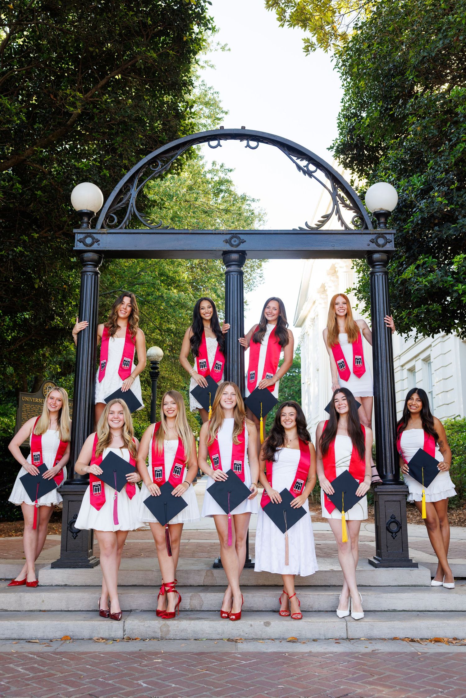 Group of female graduates in white dresses and red stoles posing under iconic arch for graduation photos.