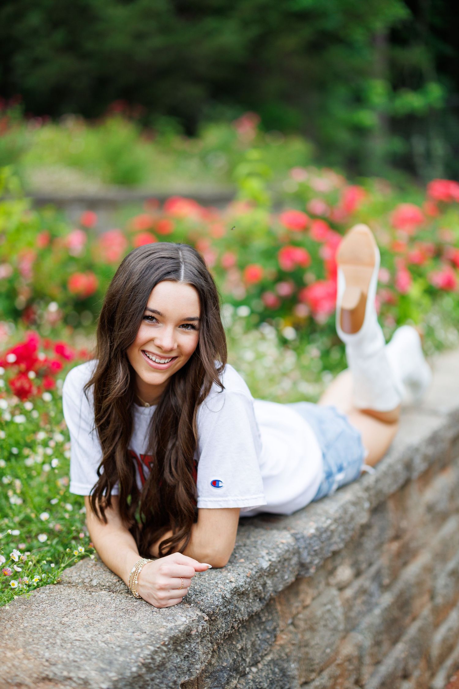 Young woman in Champion tee and denim shorts lying on stone wall smiling, with red roses blooming in background.