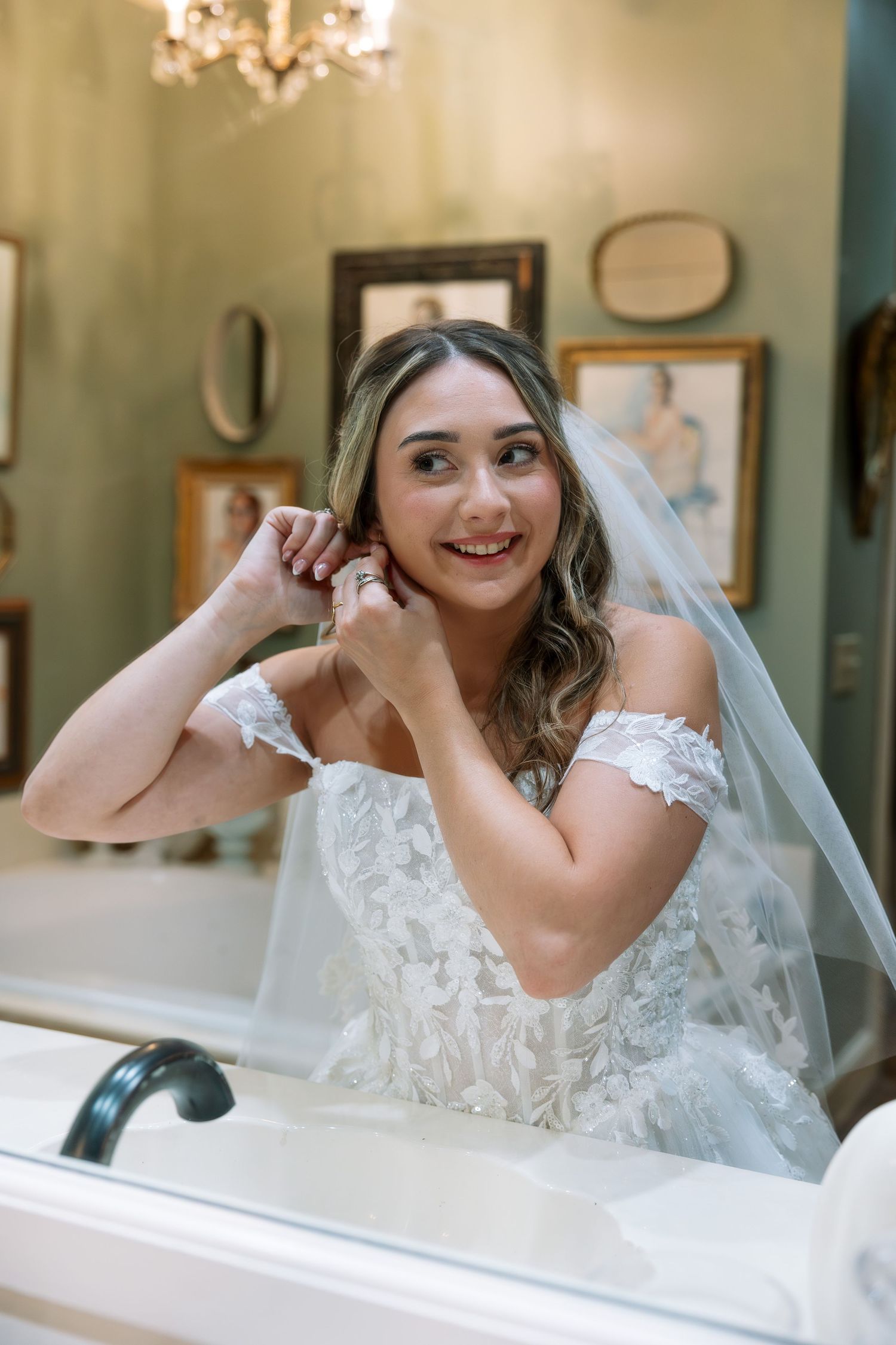 Bride in lace off-shoulder gown and veil putting on earrings, smiling in mirror with vintage decor.