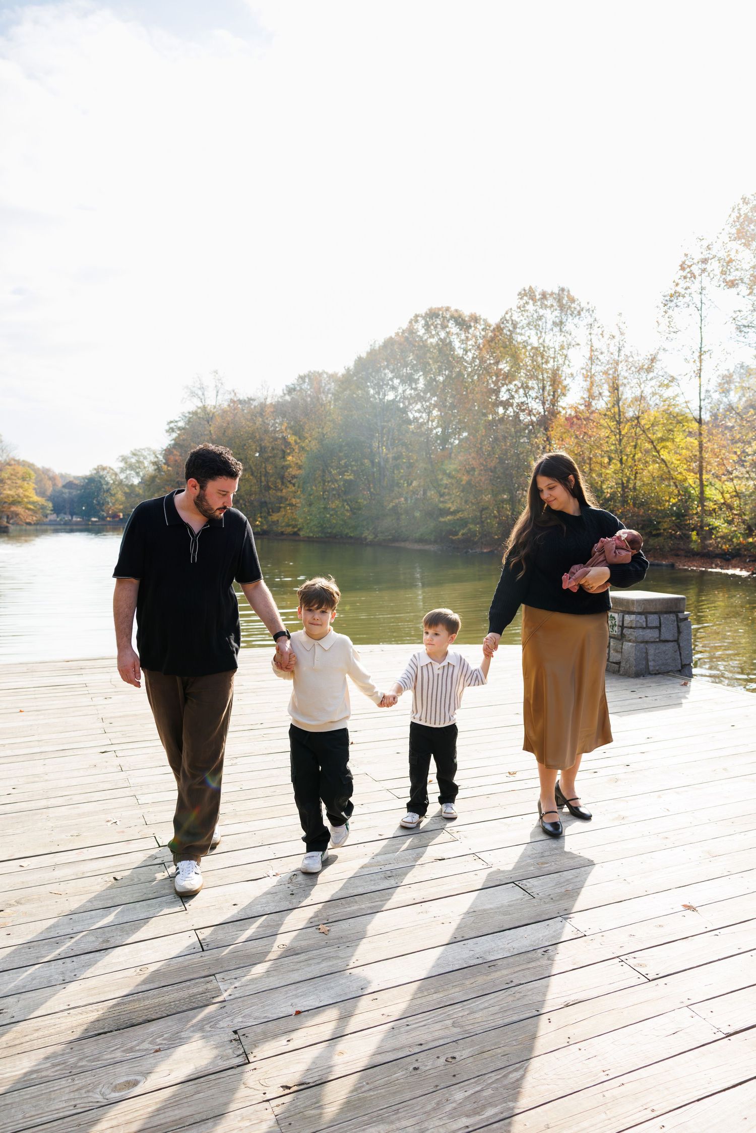 Sequence of silhouettes walking on a sunlit wooden deck by a lake during golden hour.