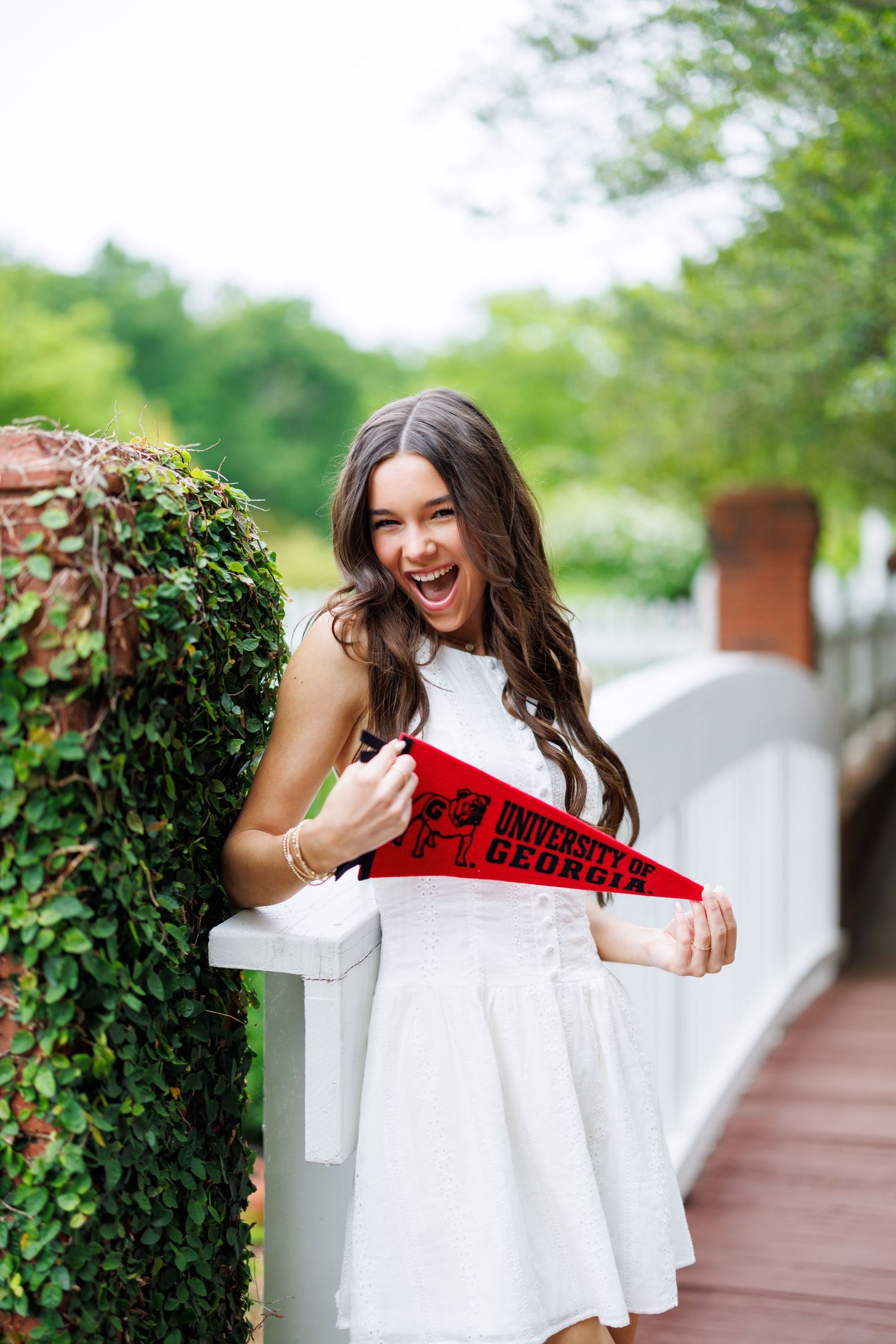 Excited young woman in white dress holding a red University of Georgia pennant on a bridge surrounded by greenery.