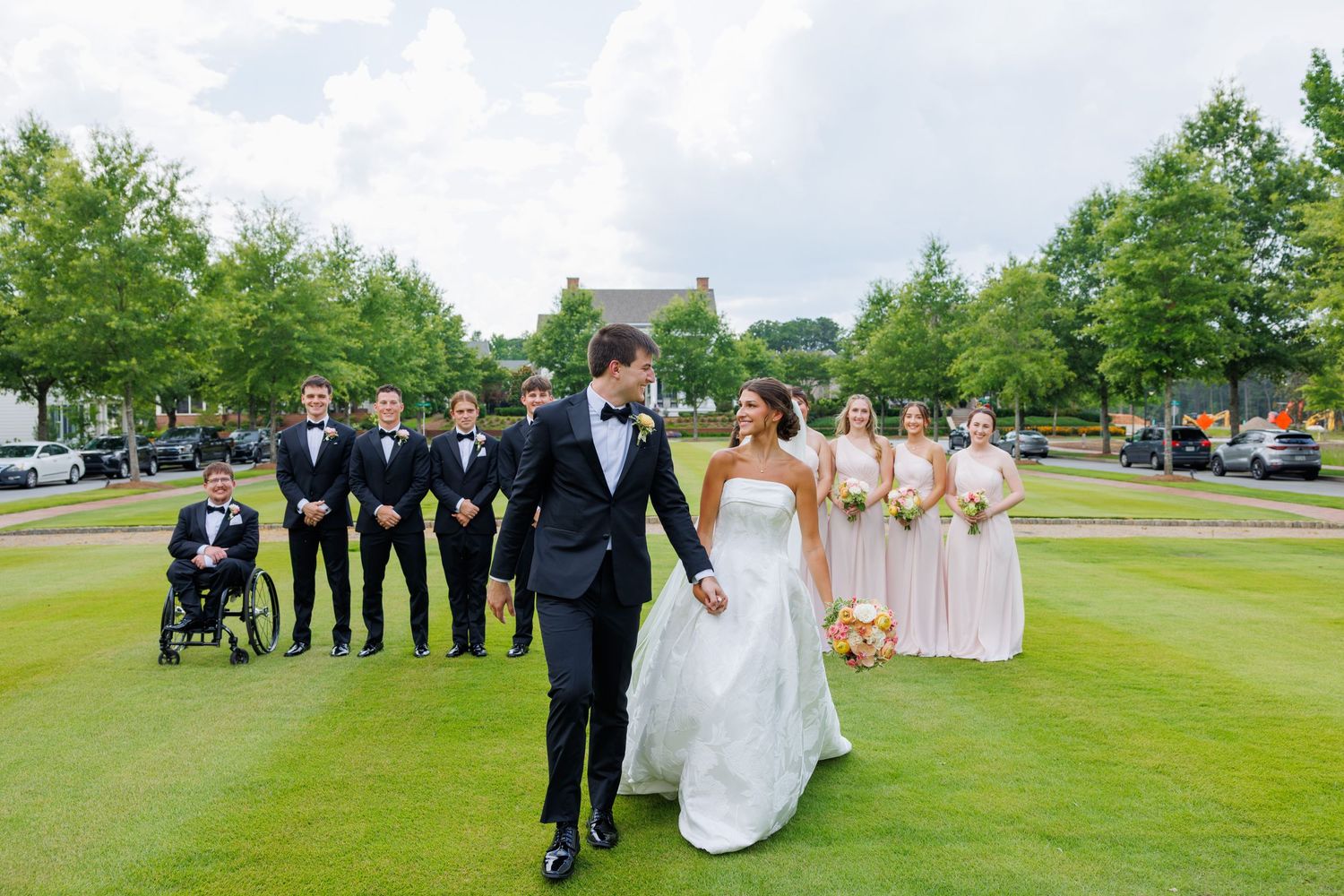 Bride and groom walk hand-in-hand on golf course with wedding party in black tuxedos and blush bridesmaid dresses behind them.
