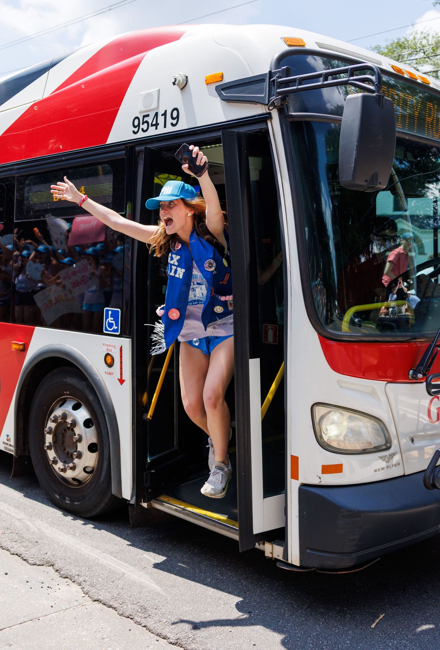 An excited young woman leans out of a red public bus door, number 95419, waving enthusiastically at a crowd outside.