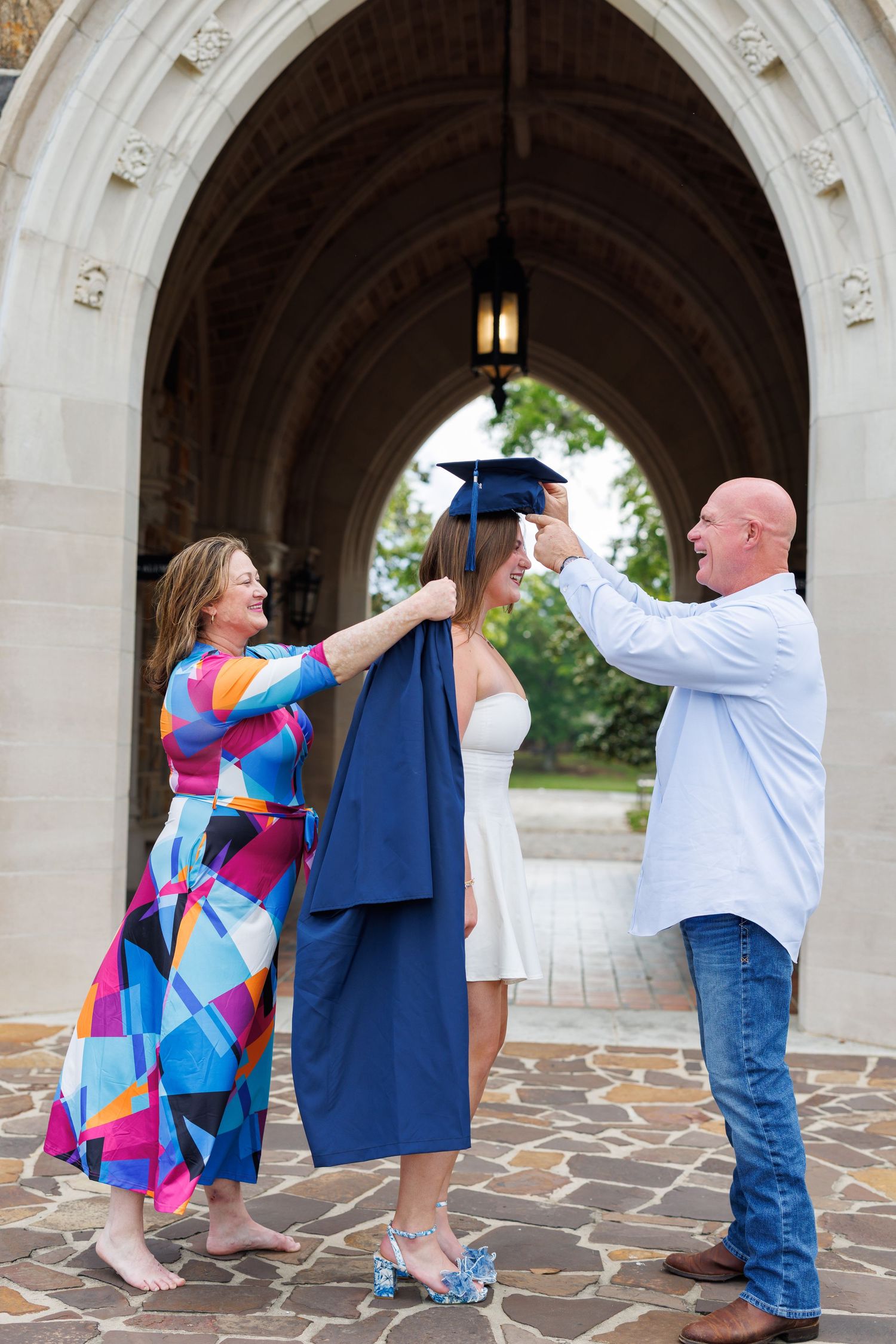 Parents helping their graduate daughter put on cap and gown under stone arch at college campus ceremony.