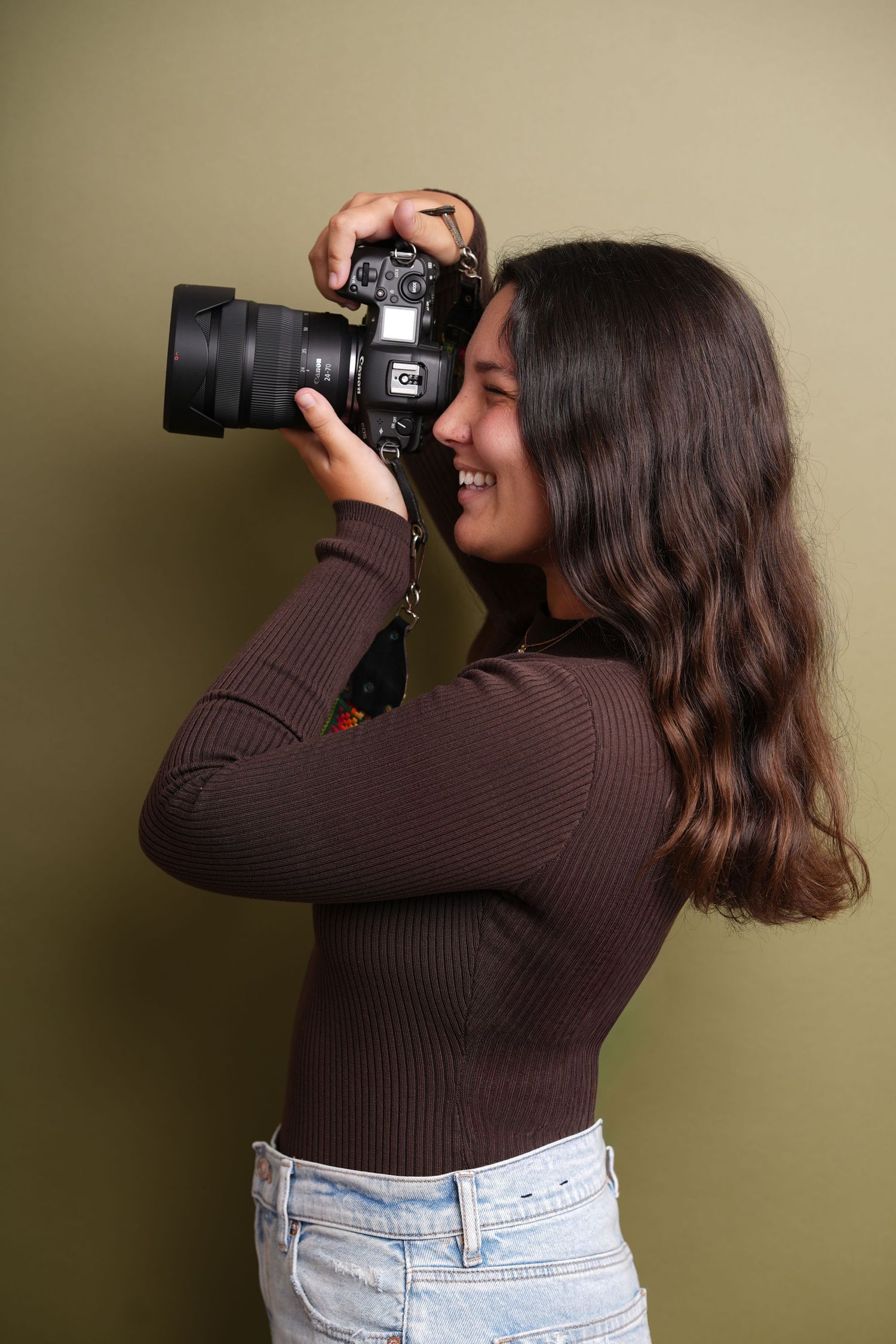 A photographer wearing a brown sweater and jeans takes photos with a professional DSLR camera against a green wall.