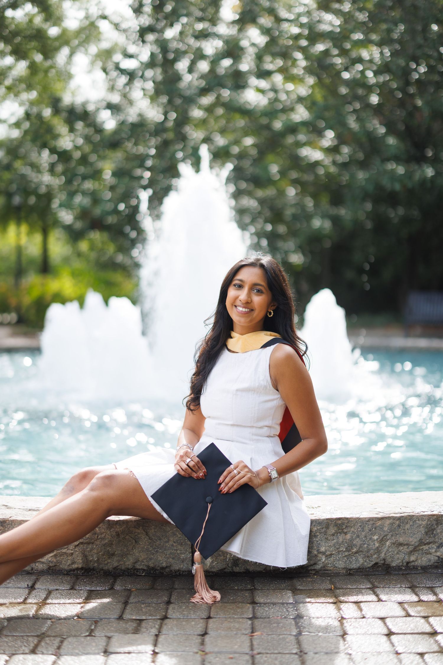 Graduate in white dress poses with diploma by fountain in outdoor garden setting.