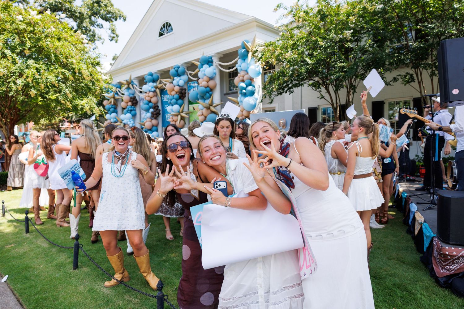 Excited college women in white dresses celebrate sorority bid day outdoors with blue balloons and confetti.